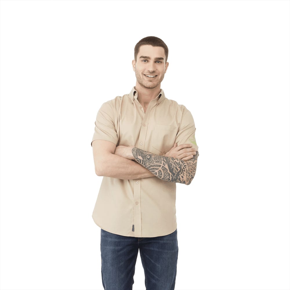 A man wearing a beige short sleeve button-up shirt with a tattoo, standing with arms crossed. The shirt has a logo. Stirling Short Sleeve Shirt - Mens - Image 1