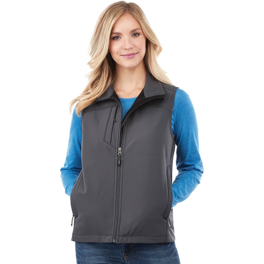 A women's softshell vest in dark grey, featuring a zip front and a logo. Model wears a blue long-sleeve top underneath. Stinson Softshell Vest - Womens - Image 1