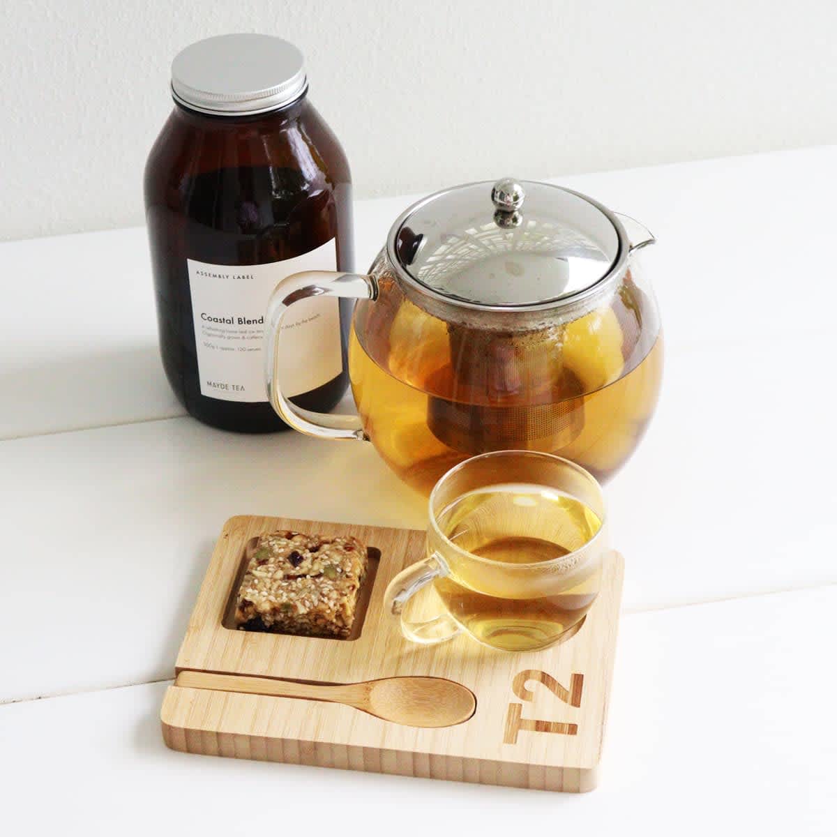 A glass teapot with a silver lid, a cup of tea, a wooden tray with a spoon, and a jar of tea leaves. Aubrey Coffee/Tea Set - Image 1