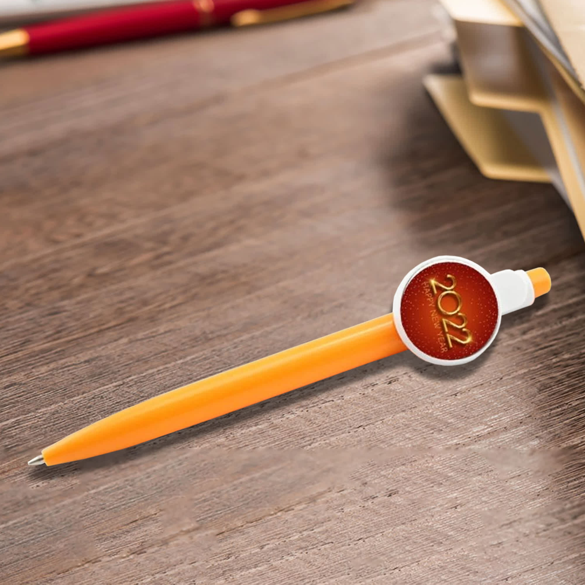 An orange plastic pen with a circular badge on the side, resting on a wooden surface beside stationery. Button Badge Pen - Image 1