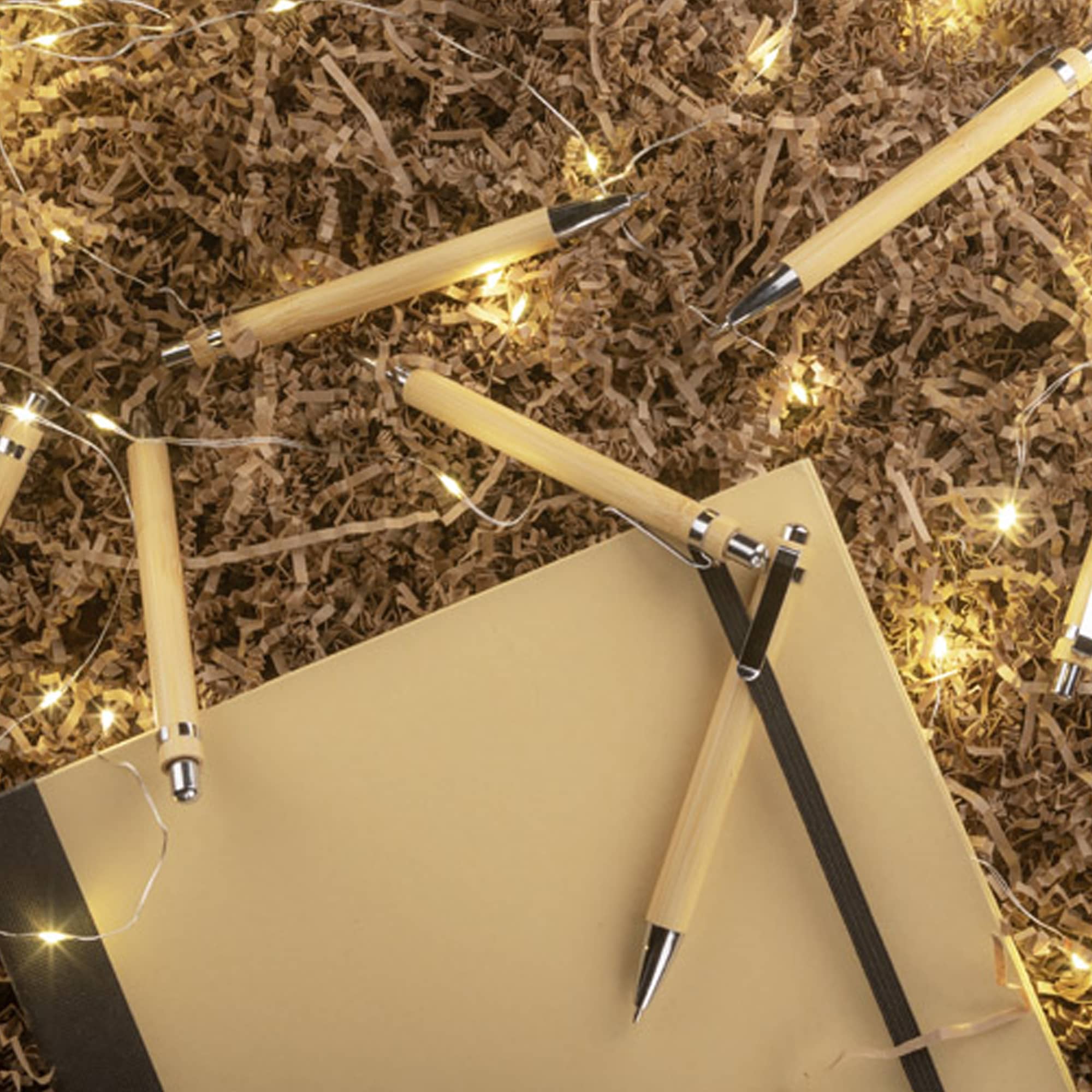 A collection of bamboo pens in natural colour scattered around a notebook and decorative lights on brown filler material. Forya Bamboo Pen - Image 1