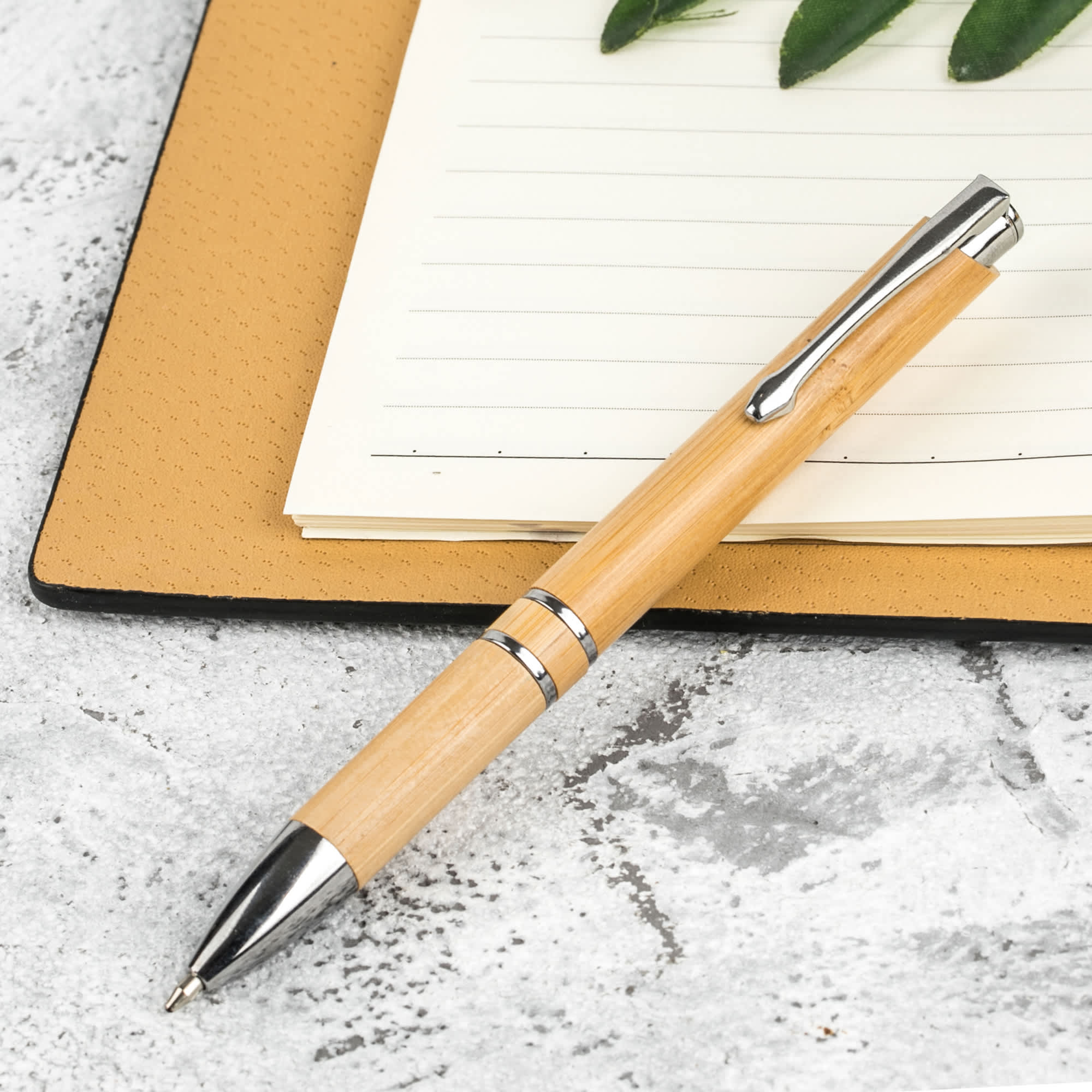 A bamboo pen with metal accents resting on a textured notebook, surrounded by greenery. Euroauz Bamboo Pen - Image 1