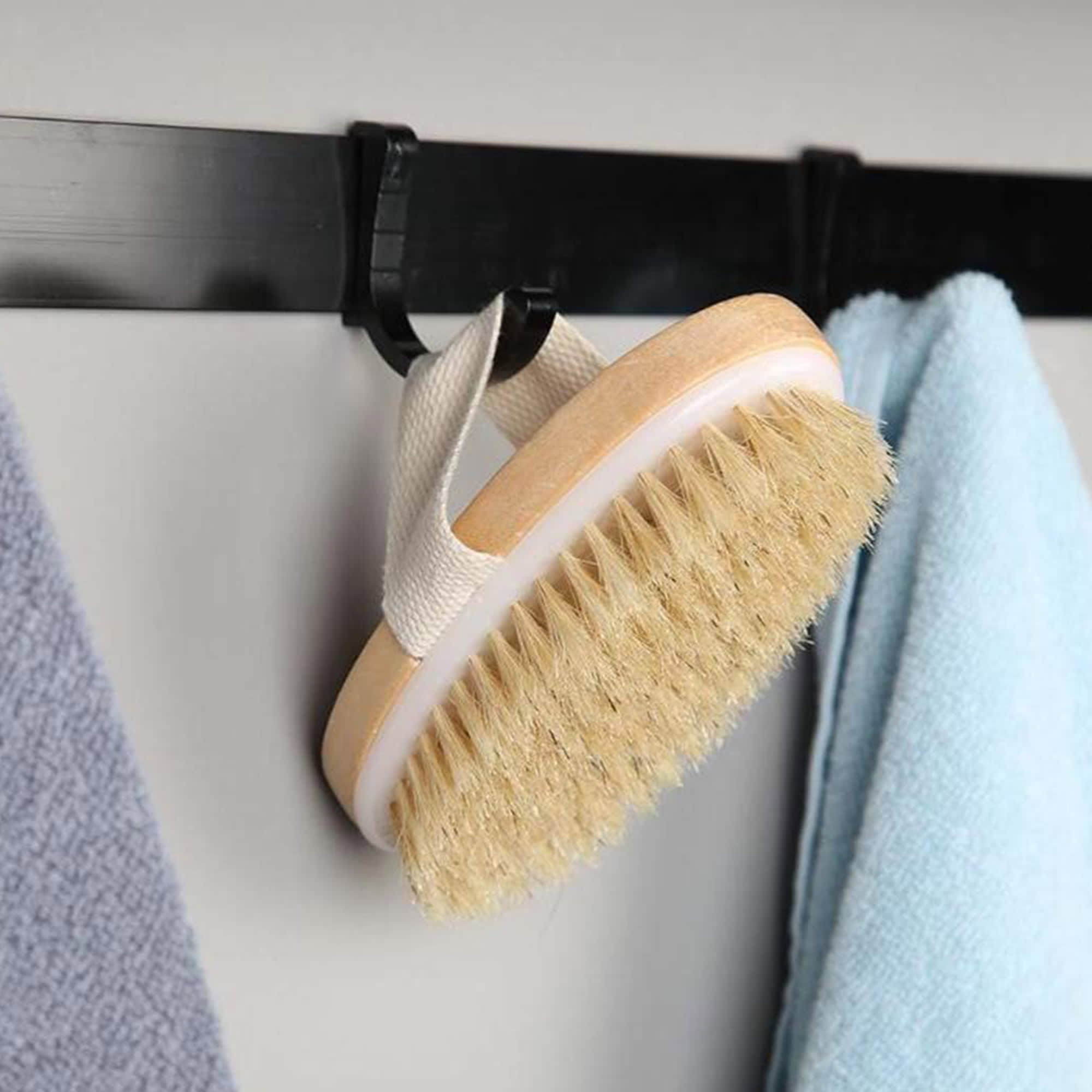 A wooden body brush with a beige bristle head and a white base, hanging on a hook beside towels. Wood Body Brush - Image 1