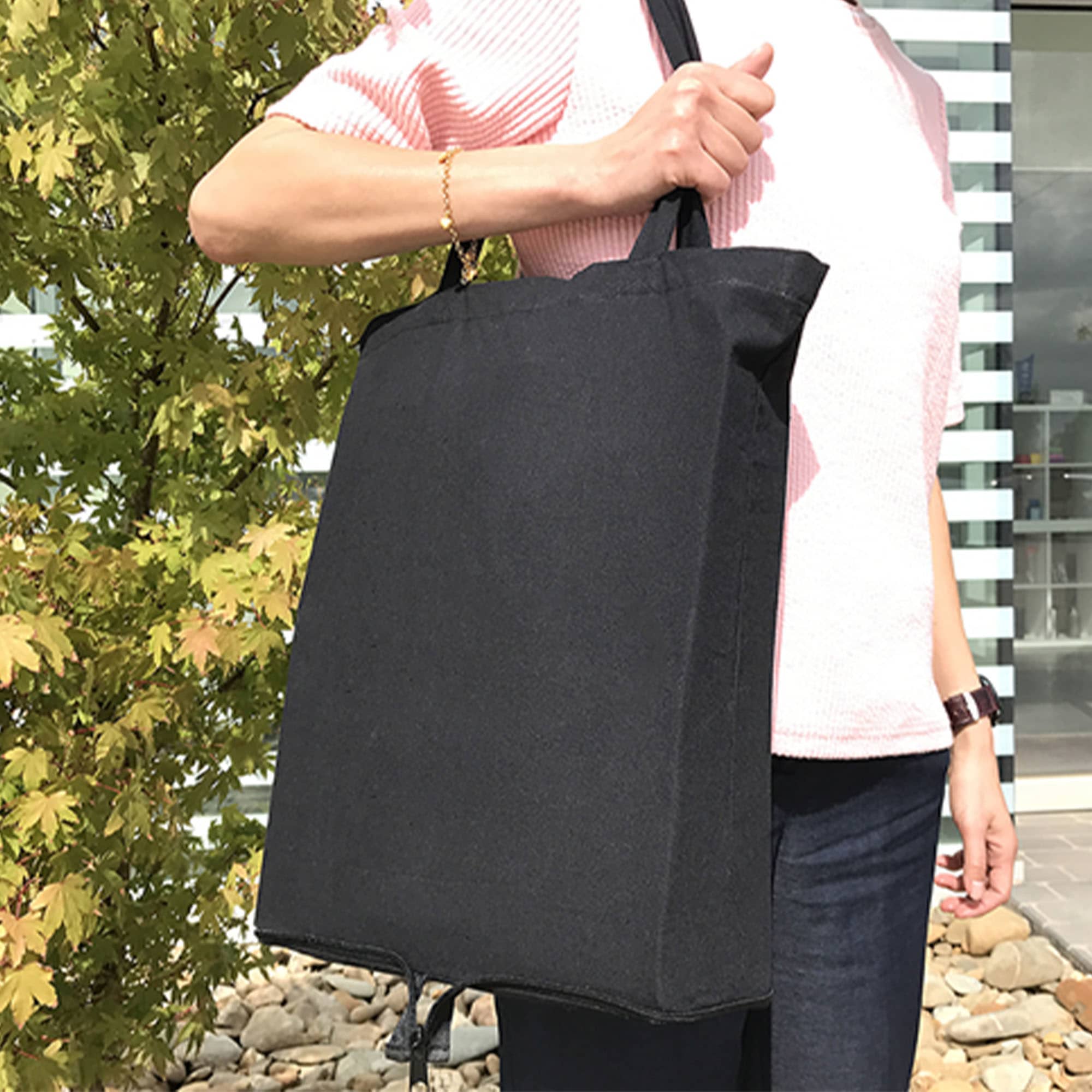 A foldable black calico tote bag with handles, held by a person against a backdrop of greenery. Foldable Calico Bag - Image 1