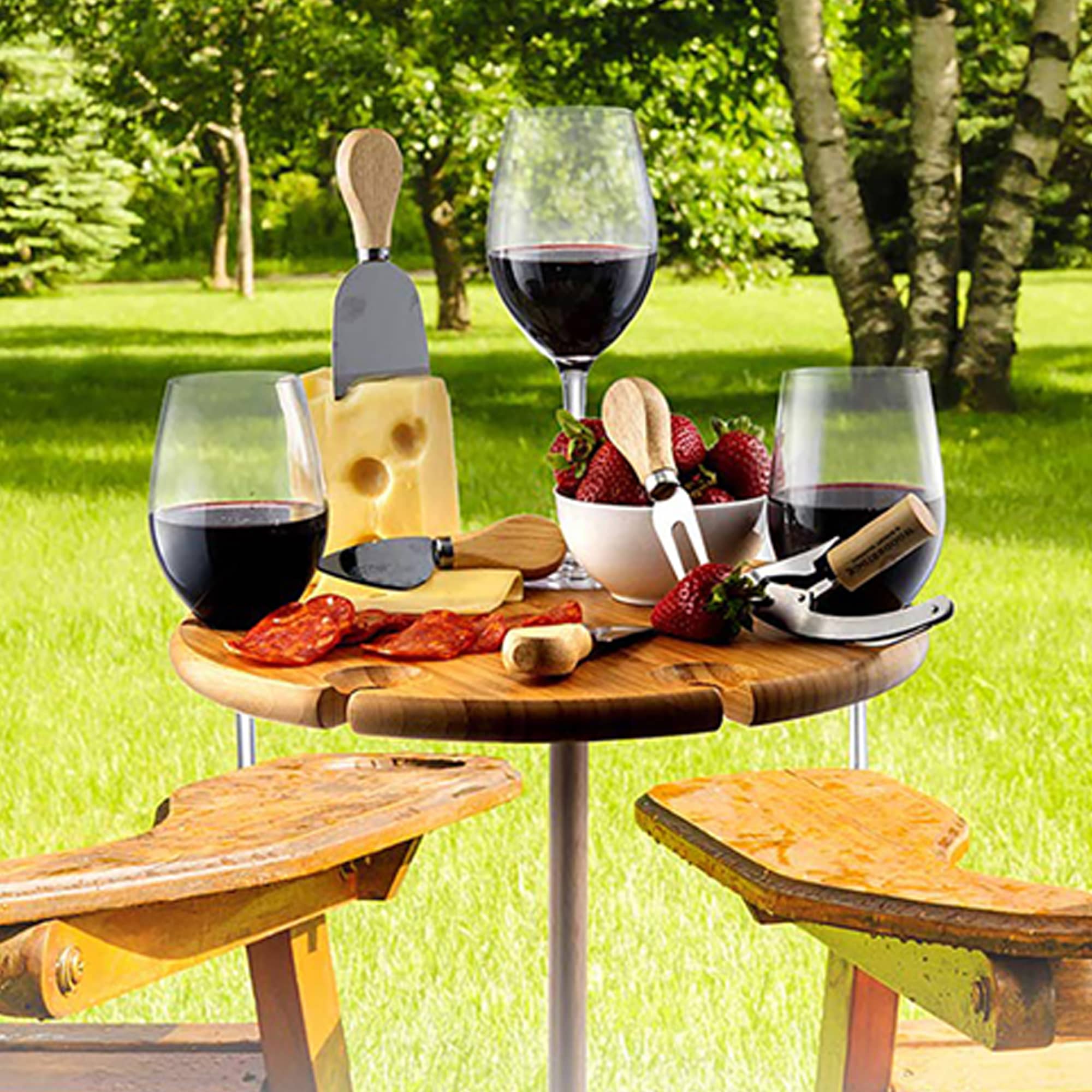 A wooden outdoor party table holds red wine glasses, cheese, strawberries, and serving utensils, set in a green garden. Bamzuki Outdoor Party Table - Image 1