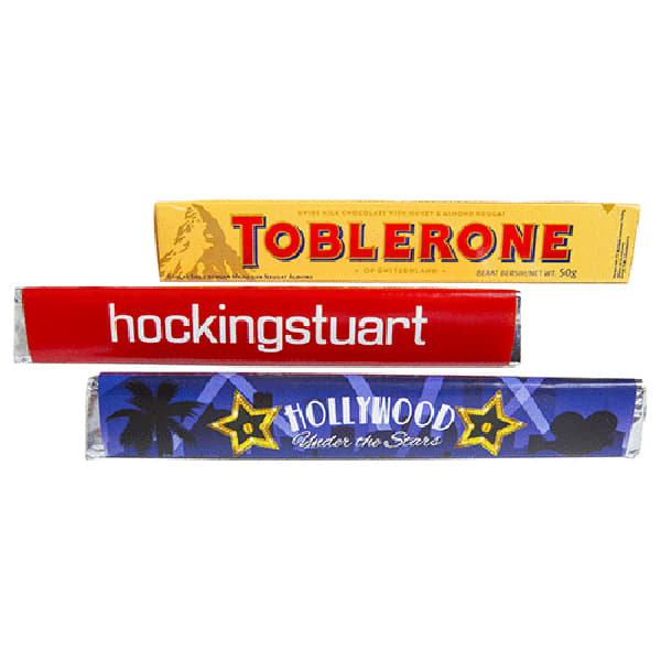 A Toblerone chocolate bar with a yellow sleeve, featuring red and blue wrapped items behind it. Toblerone 100g with Sleeve - Image 1