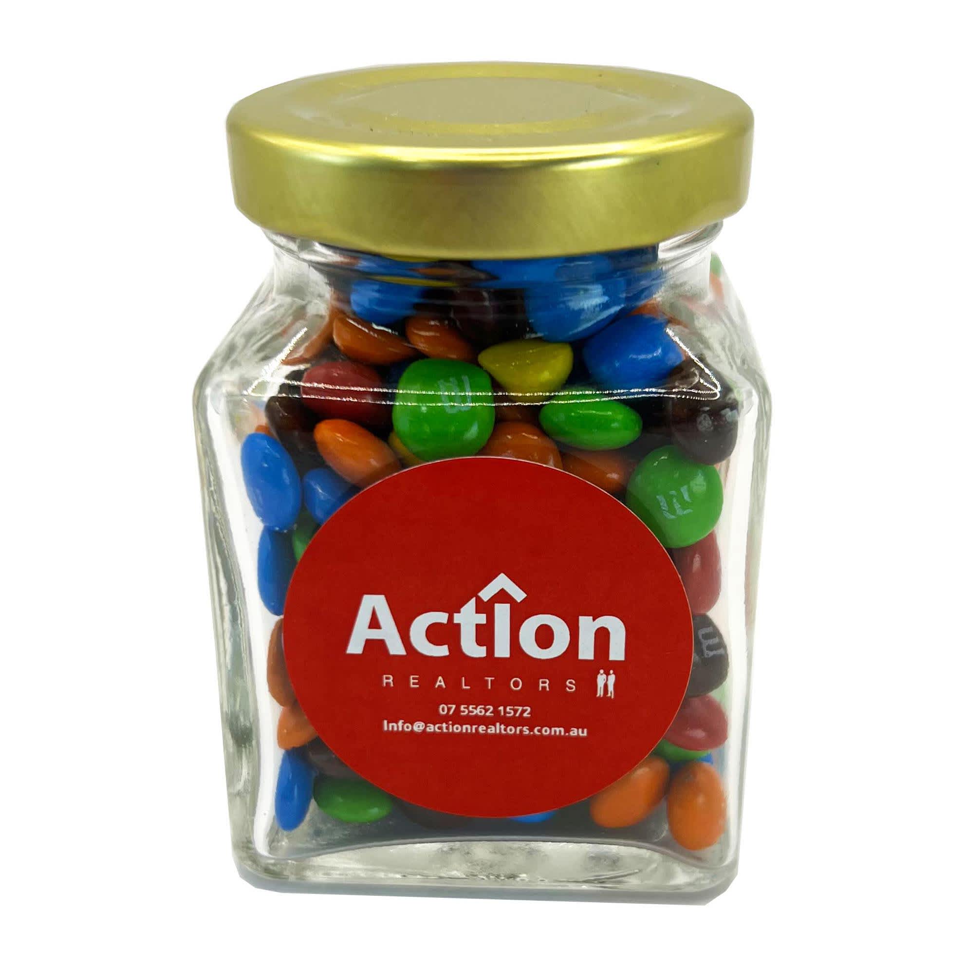 A small glass jar filled with colourful M&Ms in blue, green, red, and orange, topped with a gold lid. Small Glass Jar with Mini M&Ms 100g - Image 1