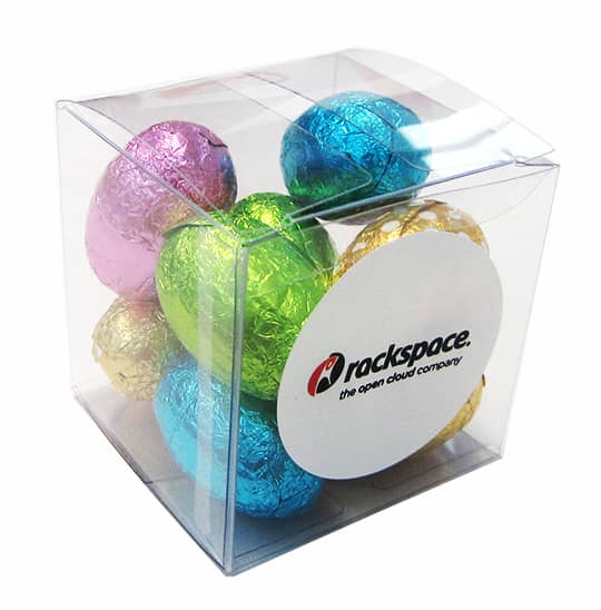 A clear cube containing mini Easter eggs in blue, pink, green, gold, and silver foil, with a logo on one side. Big Cube with Mini Easter Eggs - Image 1