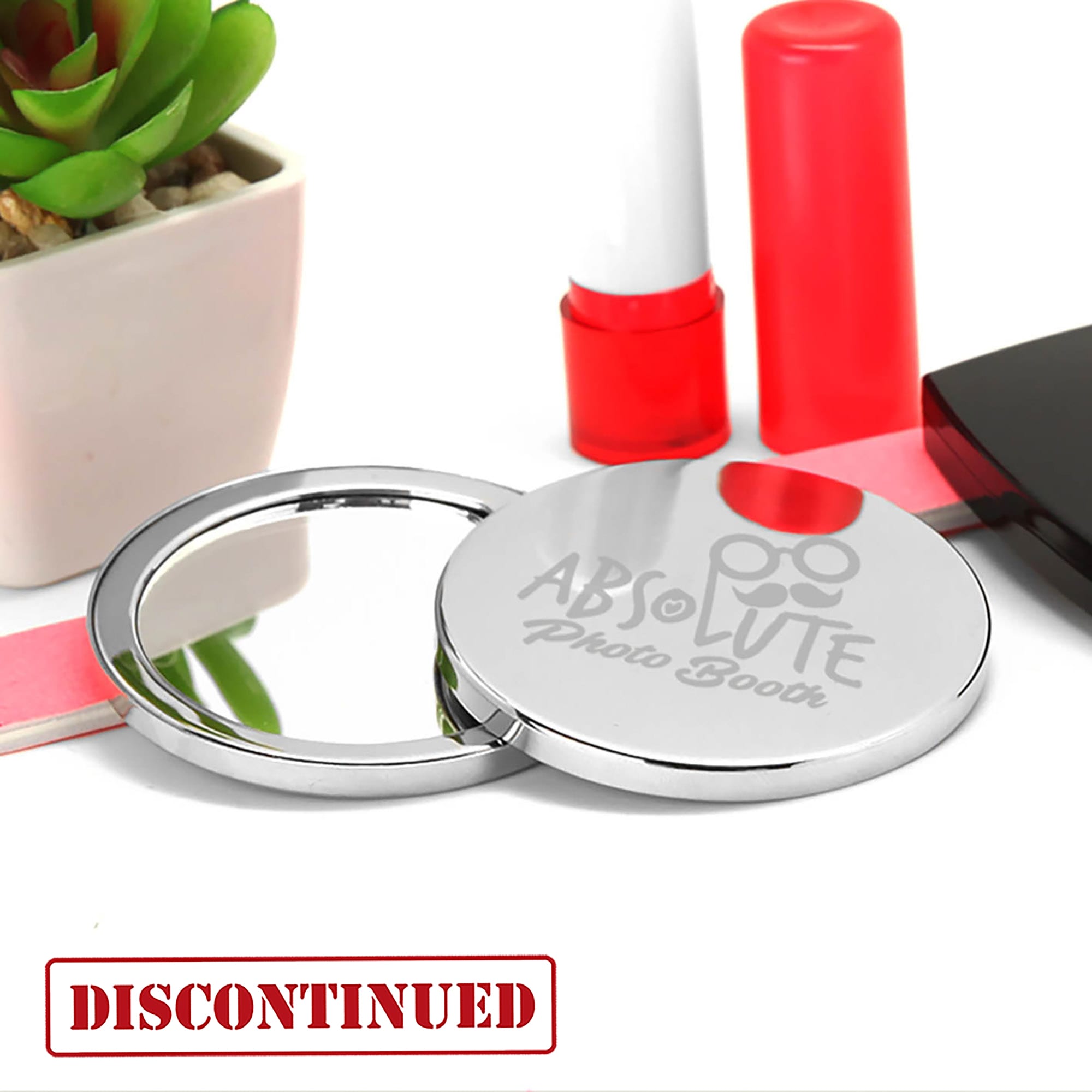 A compact mirror with a shiny silver finish, featuring a logo, placed next to a small plant and makeup items. Compact Mirror - Image 1