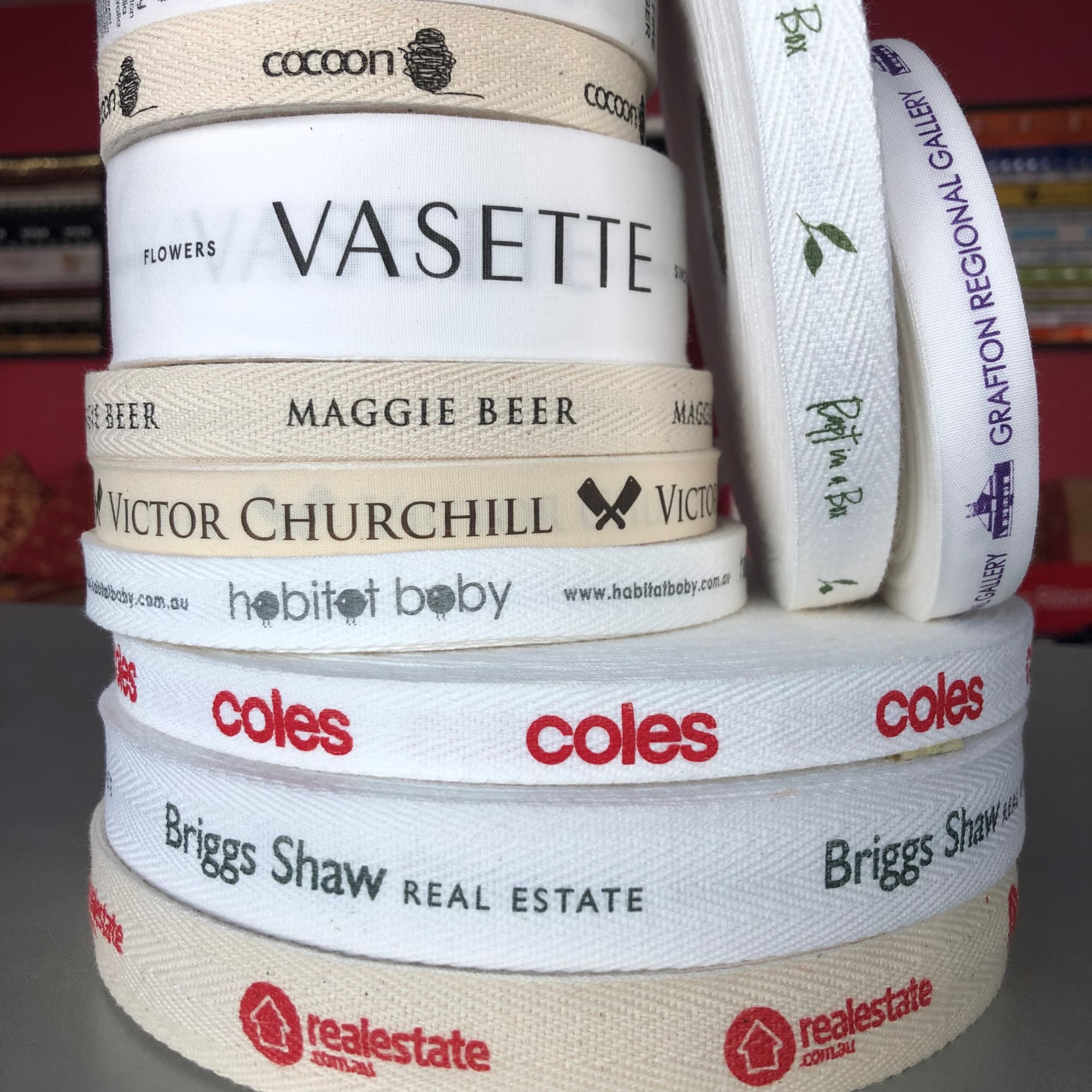A stack of custom printed flat and herringbone cotton ribbon in cream and white featuring various logos. Custom Printed Flat and Herringbone Cotton Ribbon 25mm - Image 1