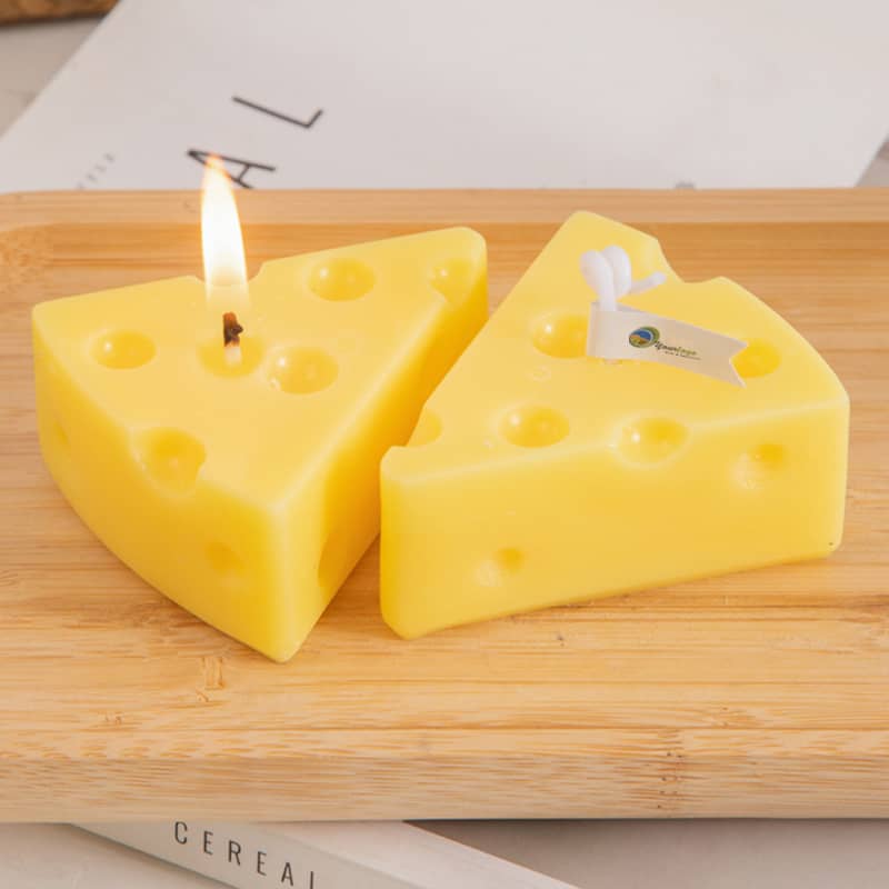 Triangle-shaped candles in yellow, resembling cheese, displayed on a wooden surface. One candle is lit. Triangle Cheese Shape Candles - Image 1