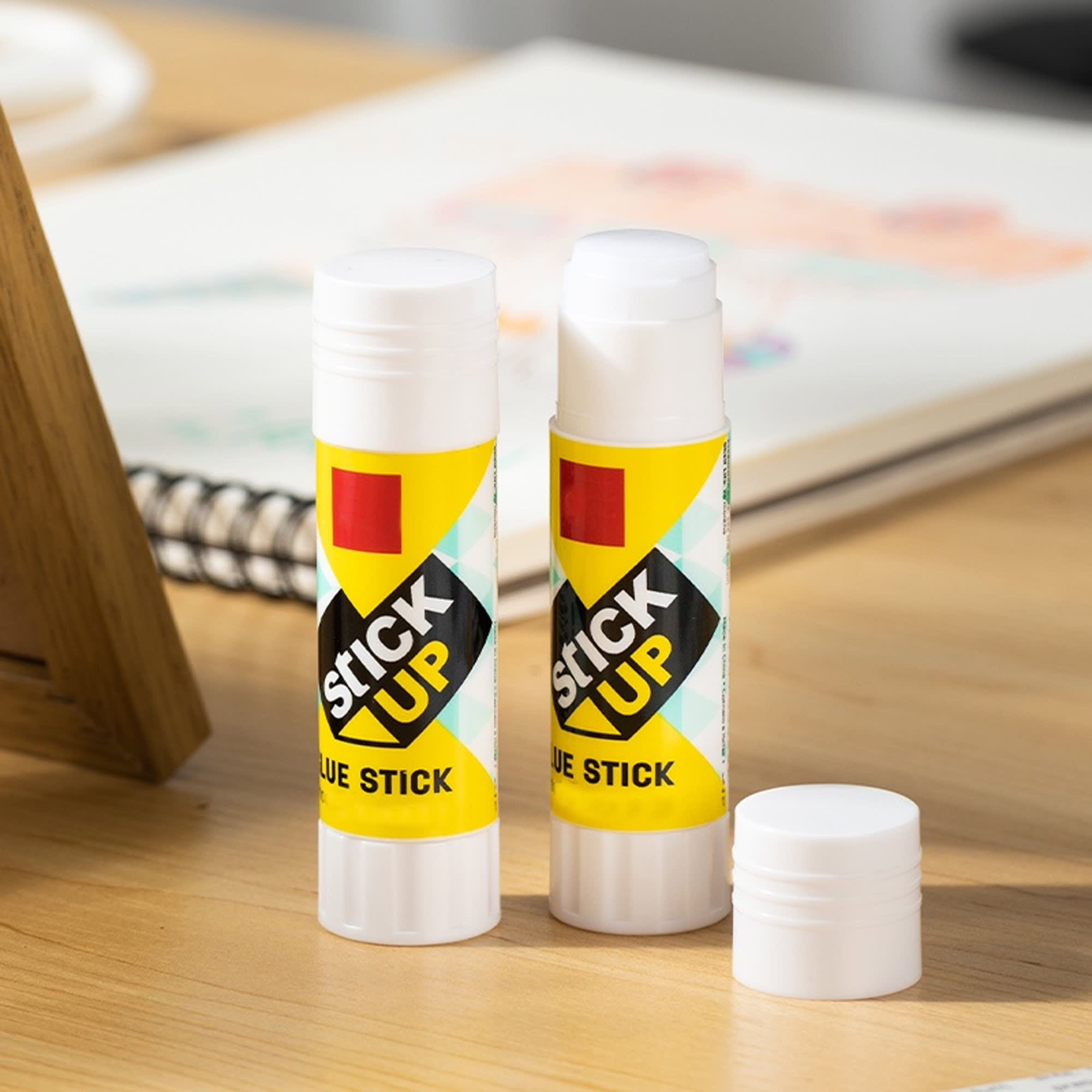 A glue stick in a bright yellow and green design, with one cap removed, placed on a wooden desk. Smart Glue Stick 9g - Image 1