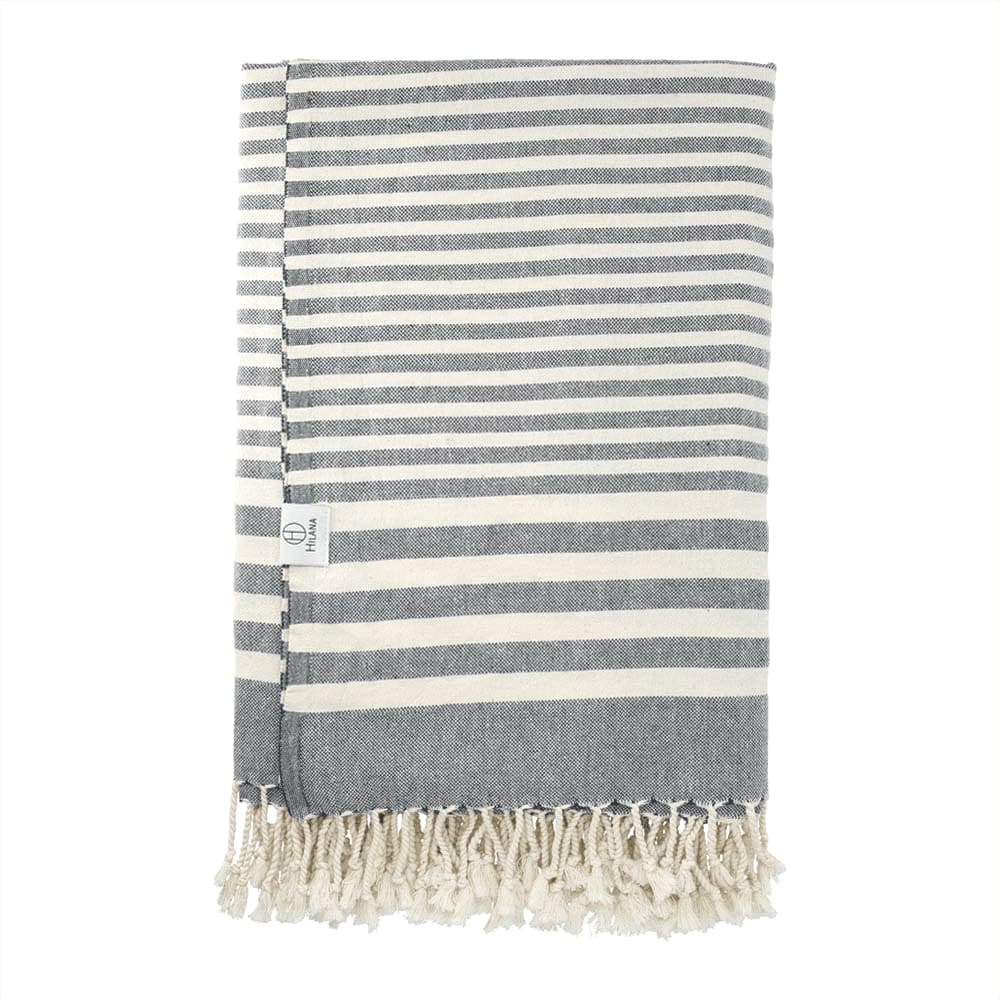 A striped throw blanket in grey and cream, featuring a fringed edge and a small logo. Hilana Upcycled Fethiye Throw Blanket - Image 1