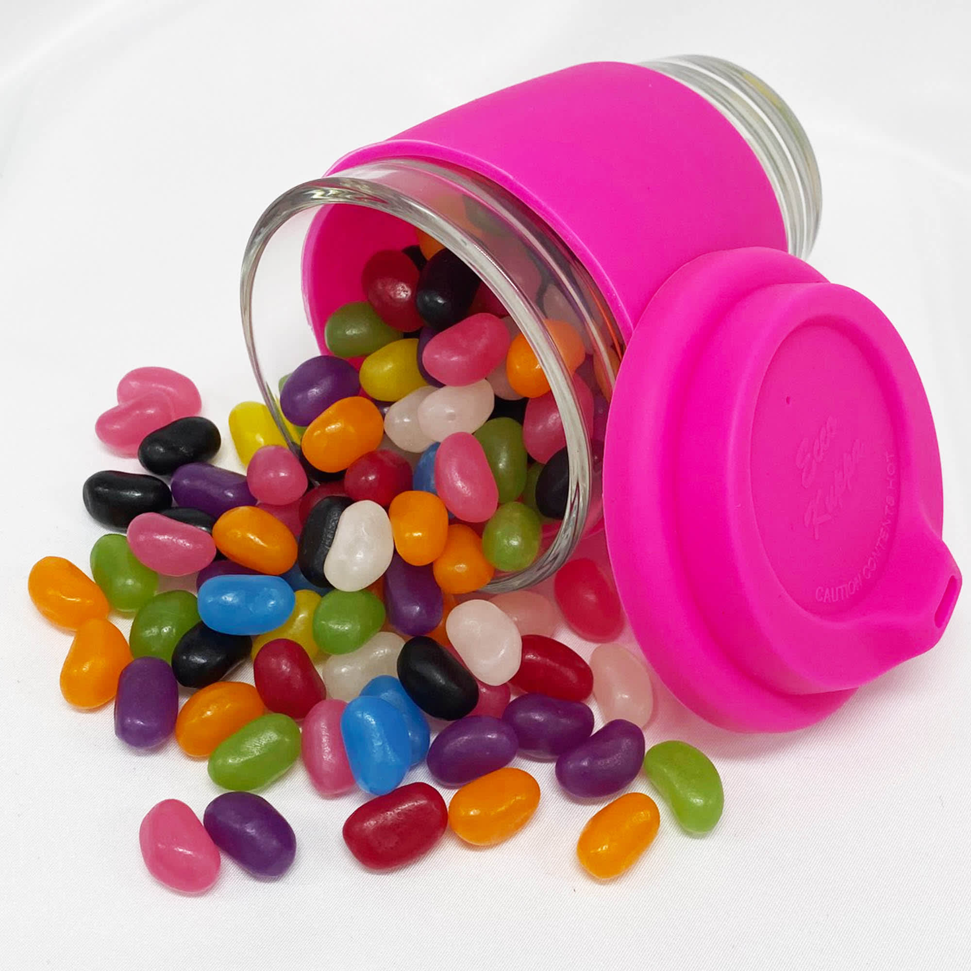 A pink silicone lid sits atop a clear glass coffee cup, filled with vibrant jelly beans in various colours scattered around. Jelly Bean In Carlo Glass Coffee Cup - Image 1