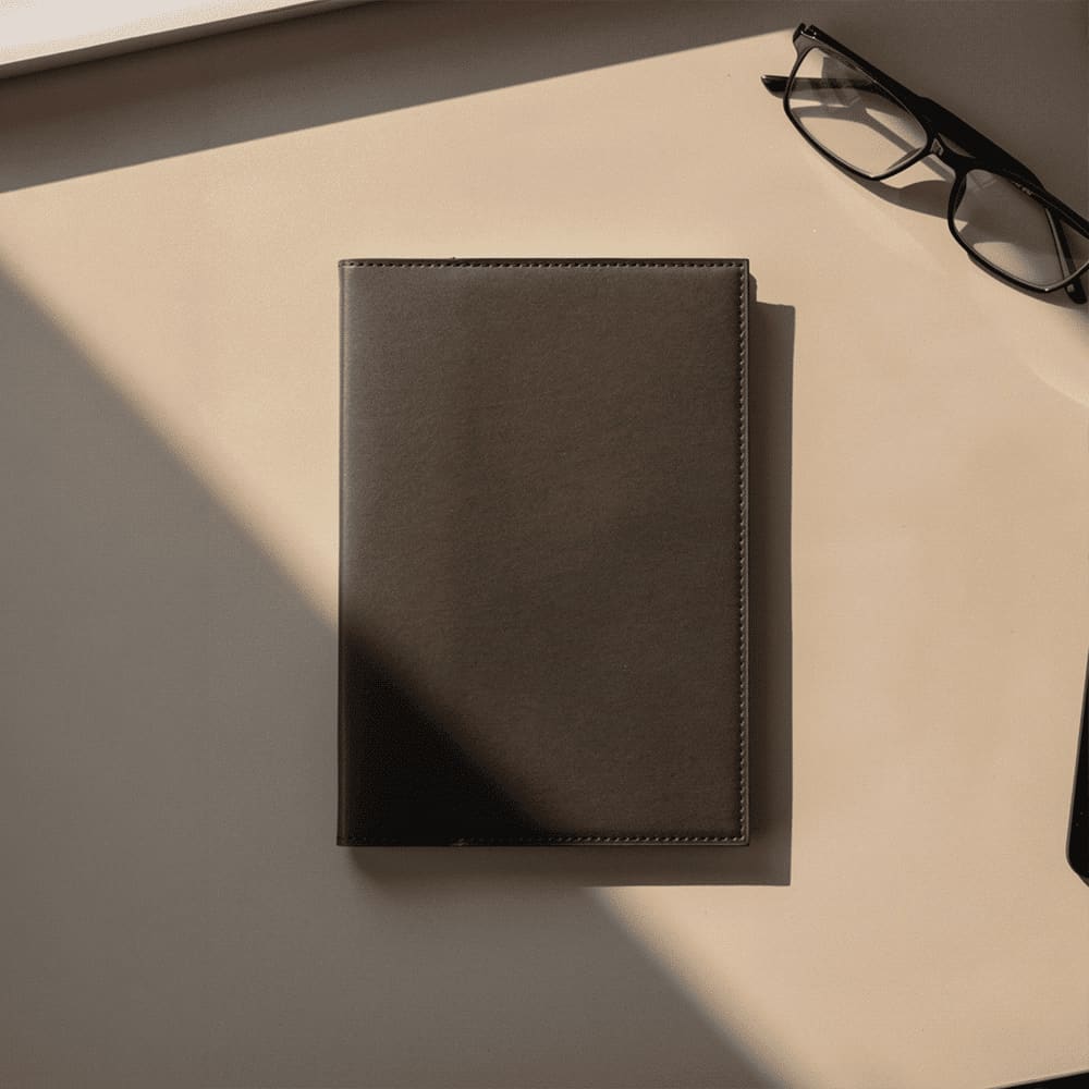A dark brown leather refillable journal lies on a beige surface, accompanied by a pair of glasses. Mela Apple Leather Refillable Journal - Image 1