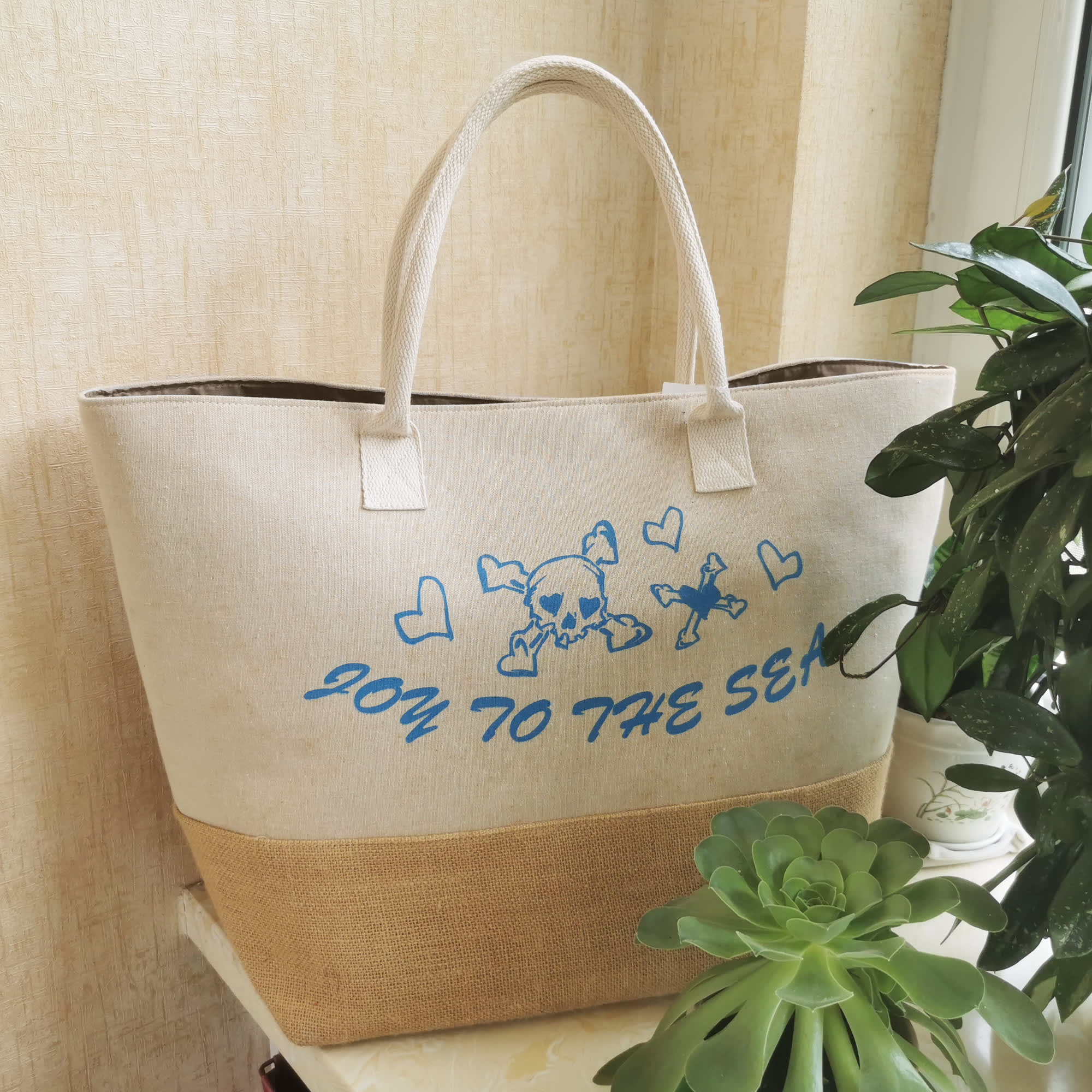 A large beige tote bag with a jute bottom and blue print design, featuring a logo and hearts. Summer Jute Bag - Image 1