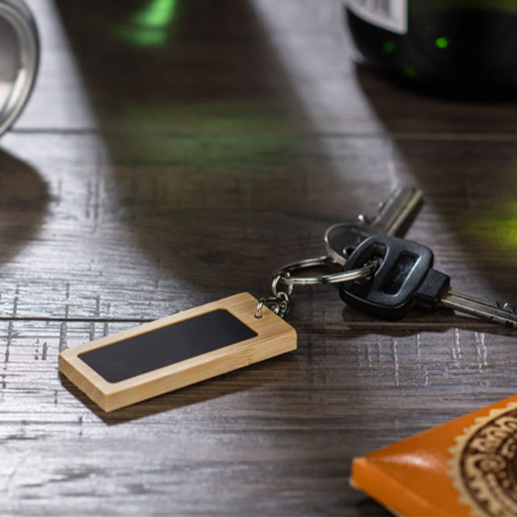 A rectangular bamboo keyring with a black surface, attached to a set of keys resting on a wooden surface. Mirage Rectangular Bamboo Keyring - Image 1