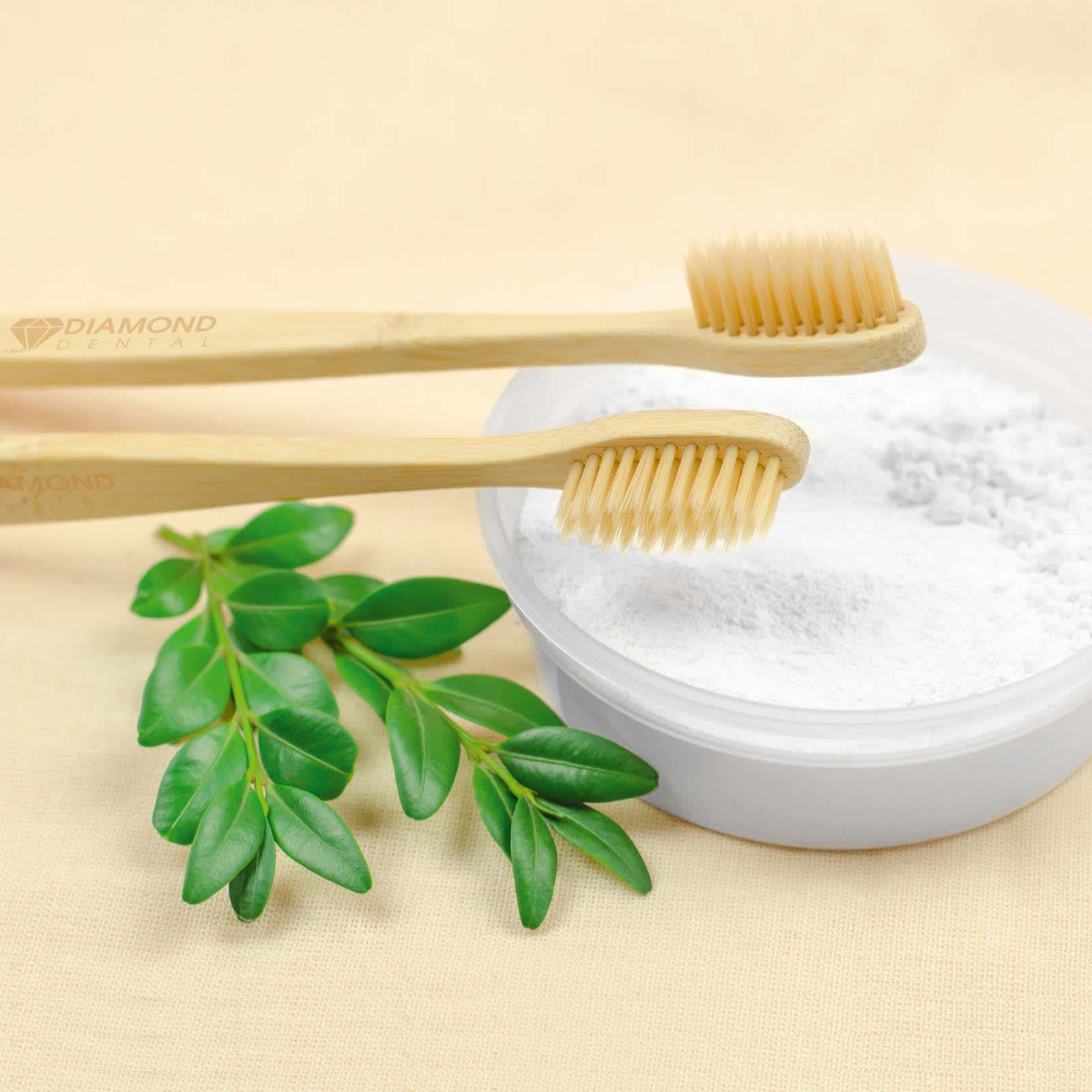 A bamboo toothbrush with soft bristles is placed next to a bowl of white powder, accompanied by green leaves. Bamboo Toothbrush - Image 1