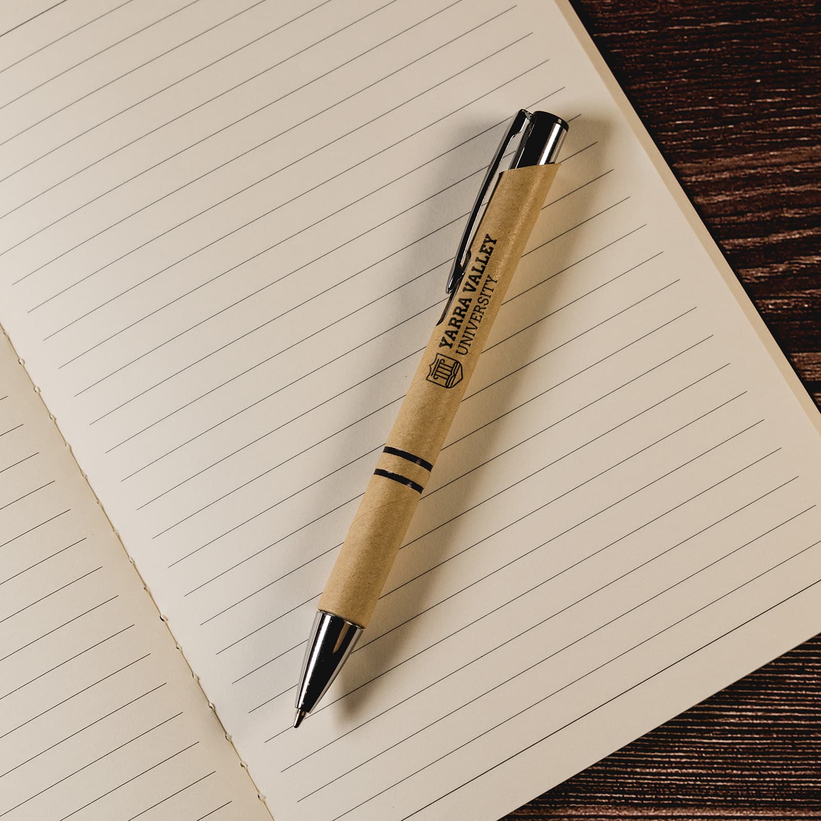 A pen with a kraft paper barrel and black accents rests atop an open lined notebook. Napier Paper Pen - Image 1