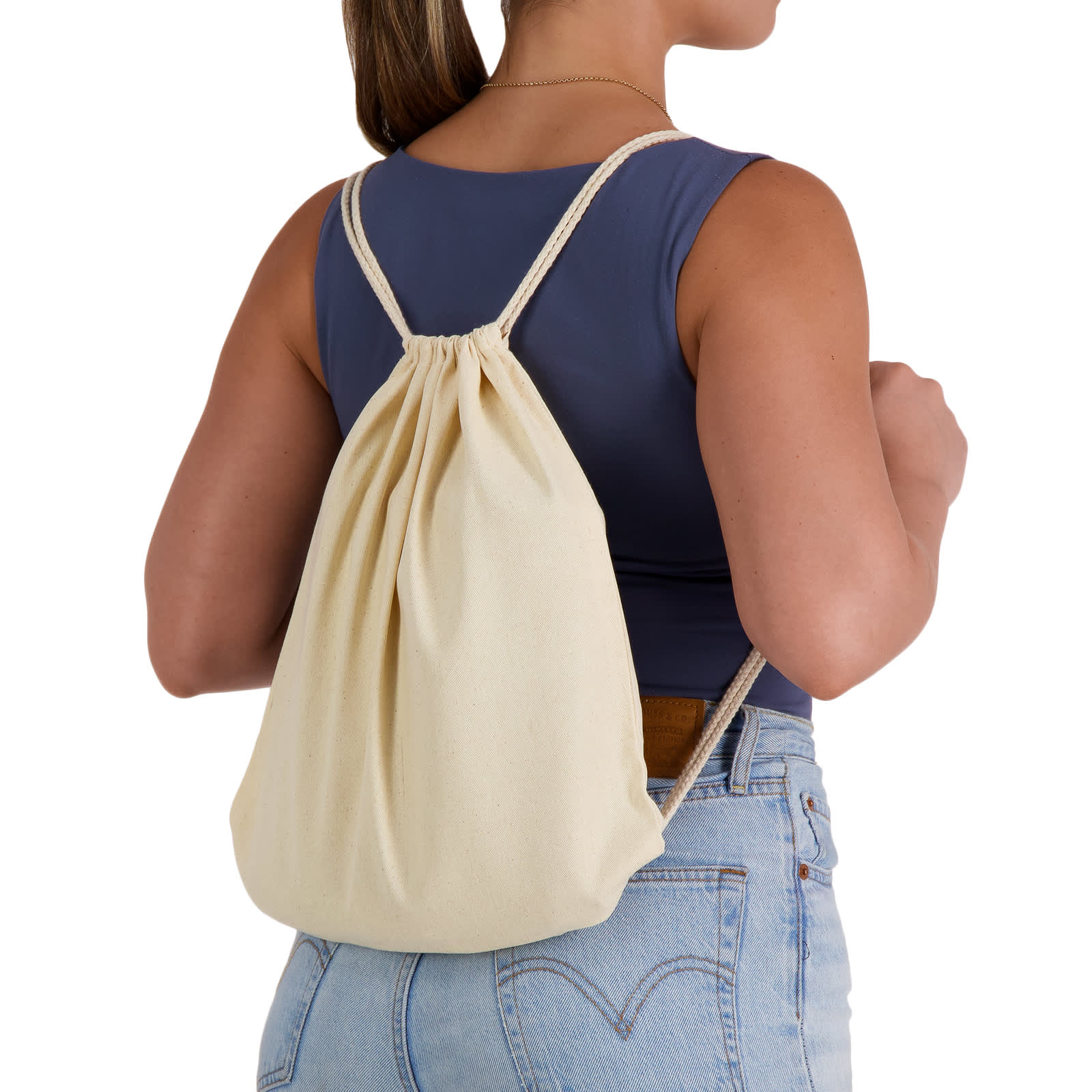 A cream-coloured drawstring backpack is worn on the back of a person, featuring simple cord straps and a gathered top. Calico Drawstring Library Backpack - Image 1