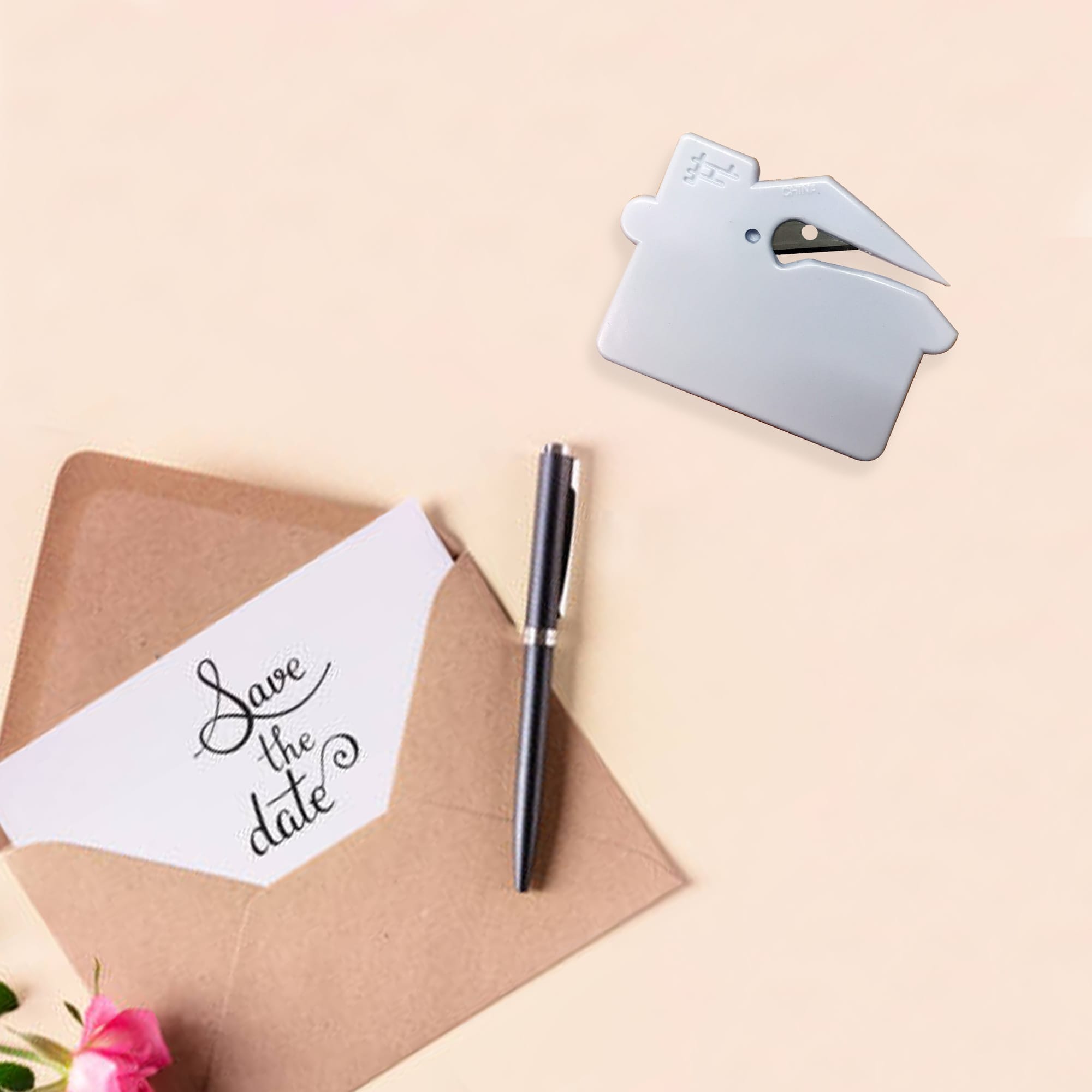 A silver house-shaped letter opener placed near an envelope and a pen, set against a soft background with flowers. House Shape Letter Opener - Image 1