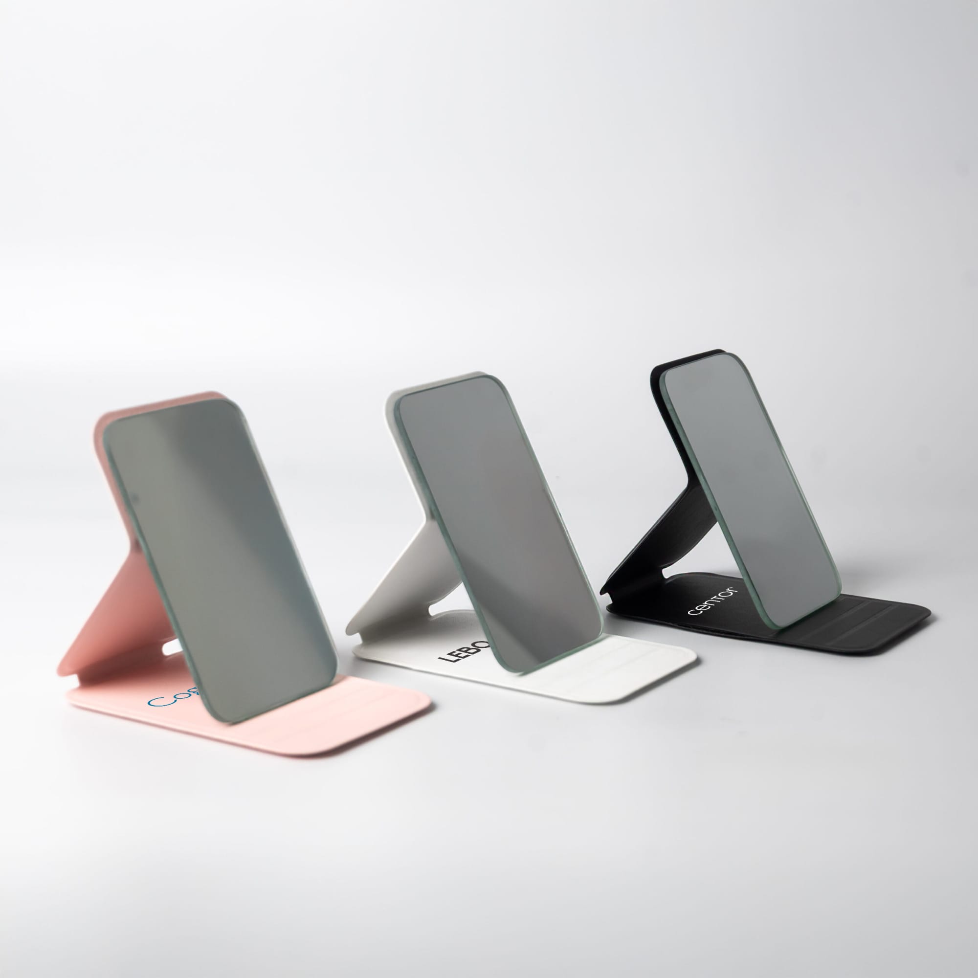 Three pocket mirrors in pink, grey, and black, each with a sleek, modern design and a stand. The Ego Pocket Mirror - Image 1
