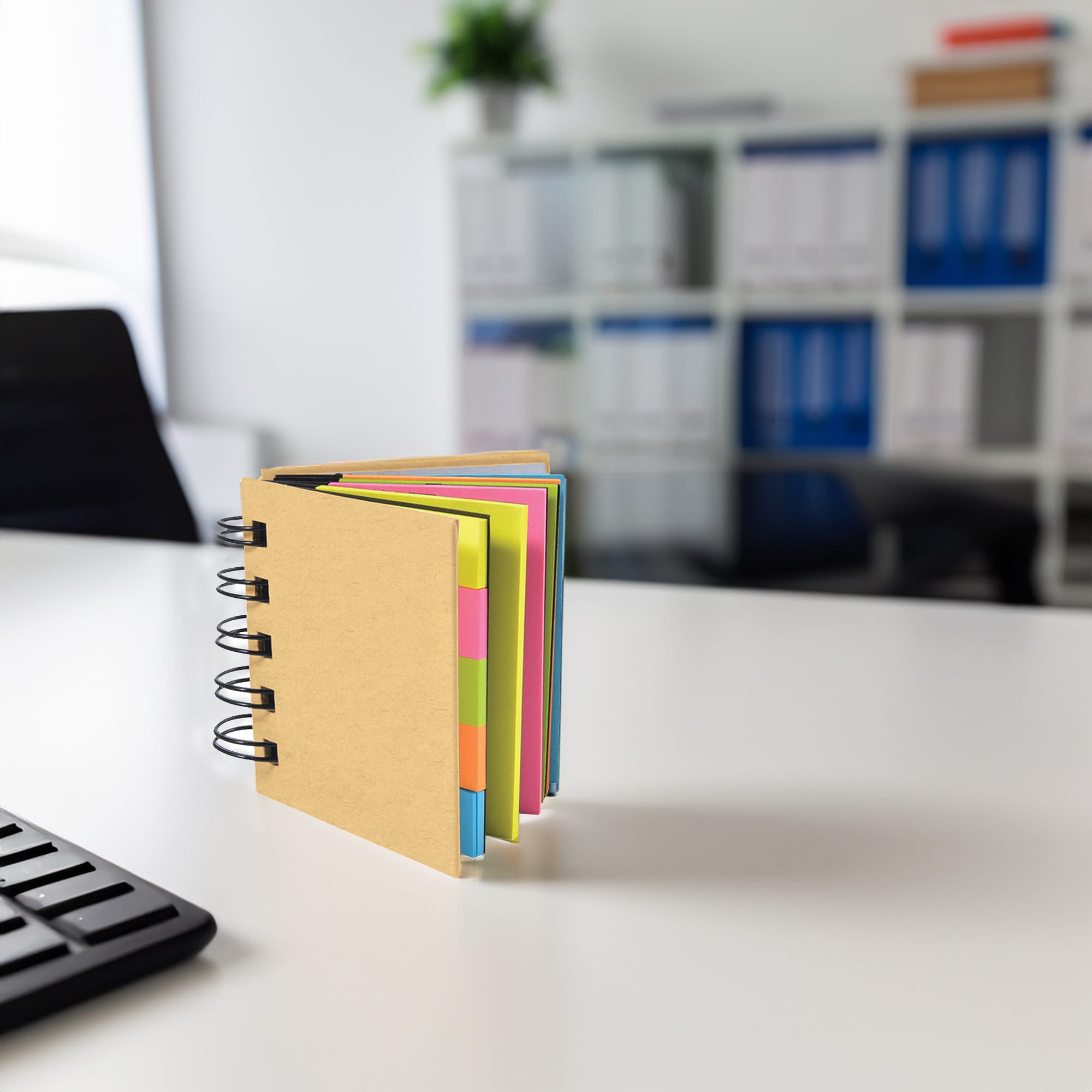 A notepad with a brown cover and colourful pages, placed on a desk in an office setting. It has a spiral binding. Jadex Note Pad - Image 1