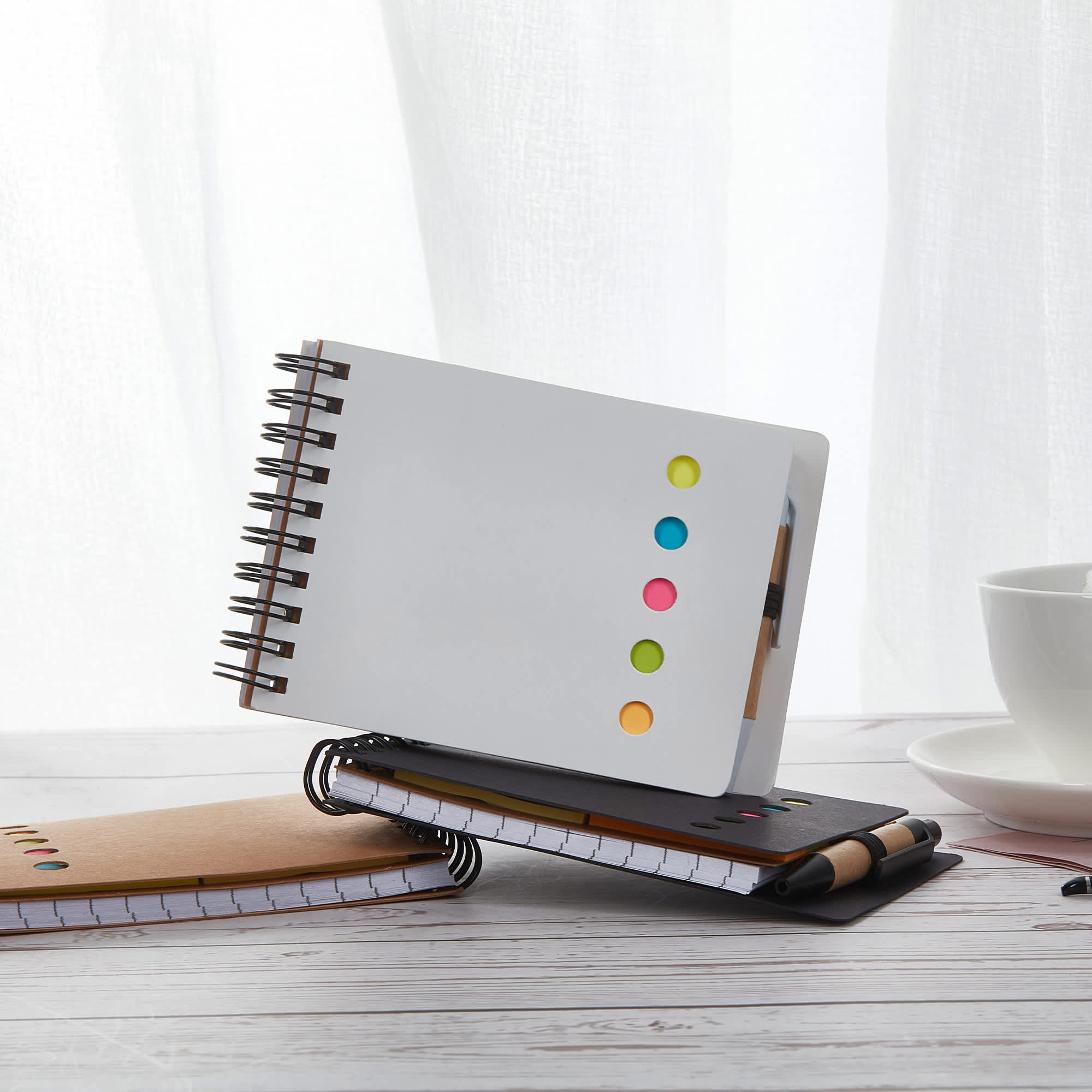 A white spiral-bound note pad with colourful sticky dots, placed on a wooden table alongside two other notebooks. Jotty Sticky Note Pad - Image 1