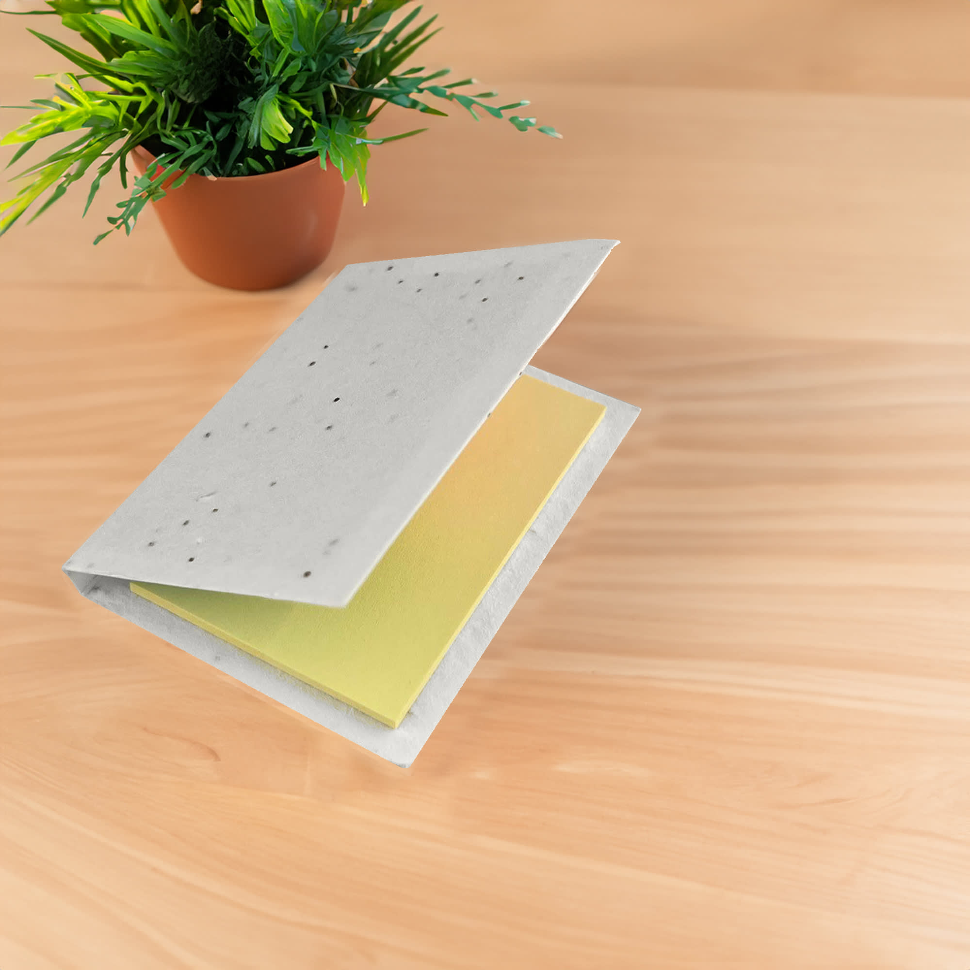 A sticky note pad with a grey cover and yellow sticky notes inside, placed on a wooden surface beside a potted plant. Corn Poppy Seed Sticky Note Pad - Image 1