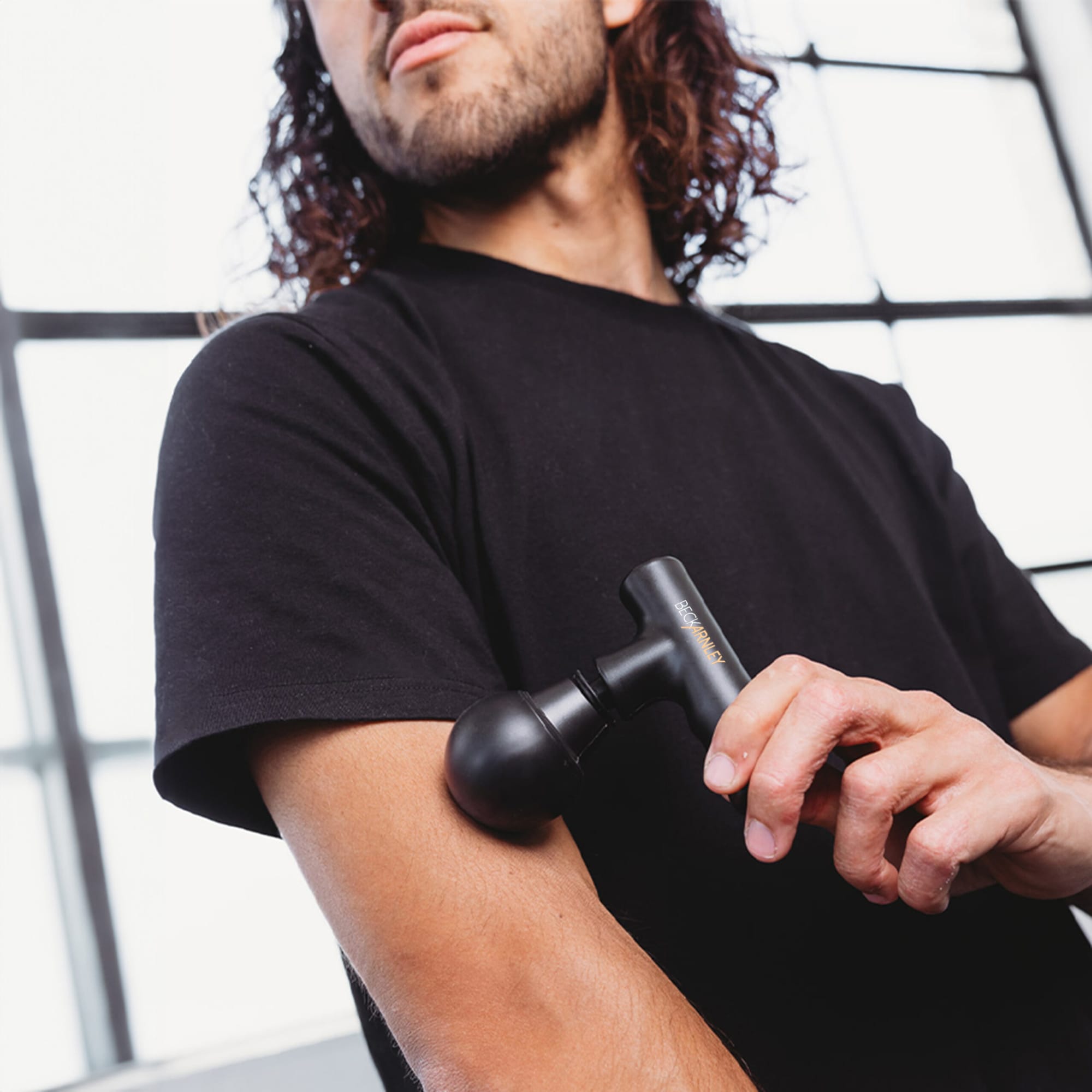 A black mini massage gun is being used on a person's arm. The setting features large windows with natural light. Mini Massage Gun - Image 1
