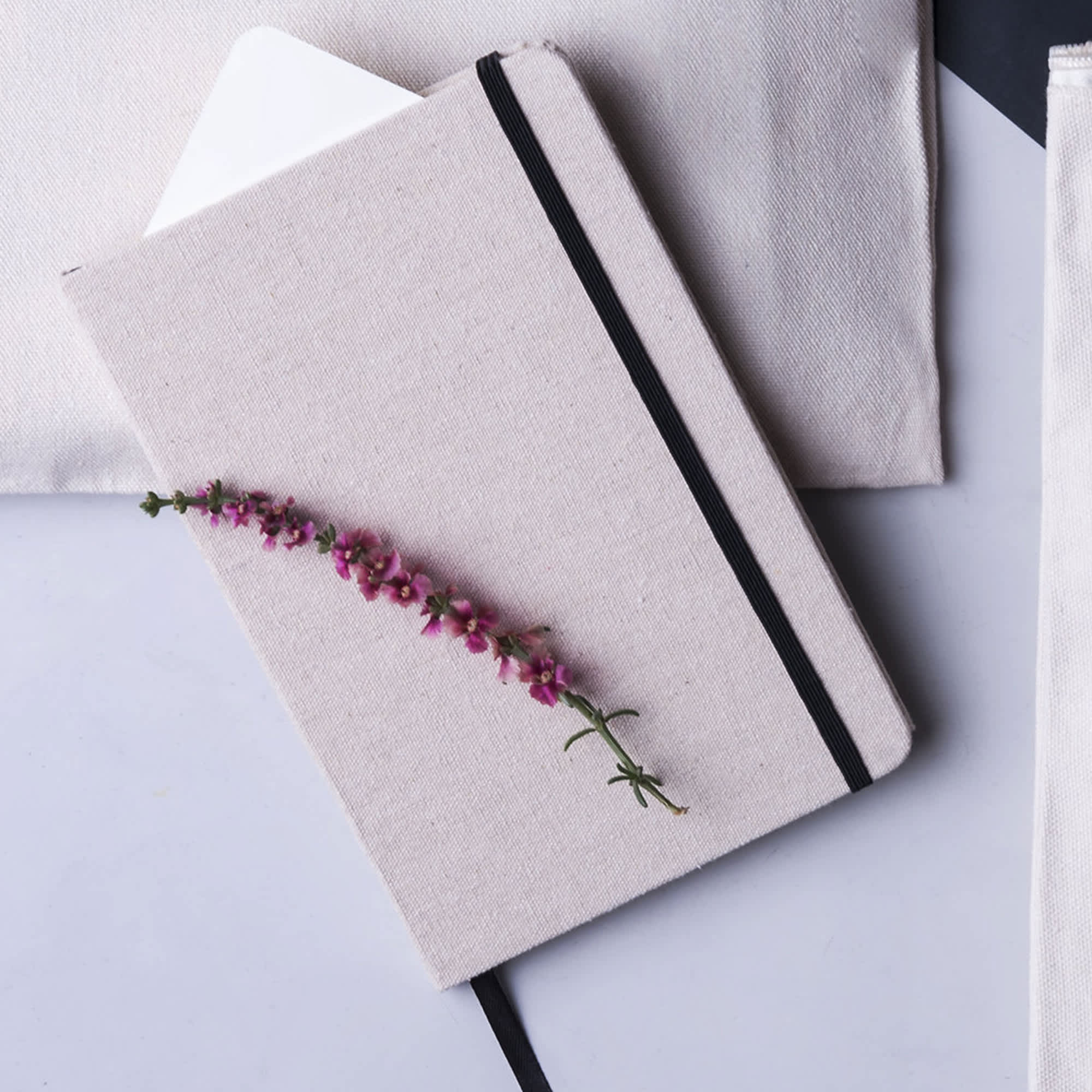 A beige A5 cotton notebook with a black elastic strap and a pink flower embellishment on the cover. Farati A5 Cotton Notebook - Image 1