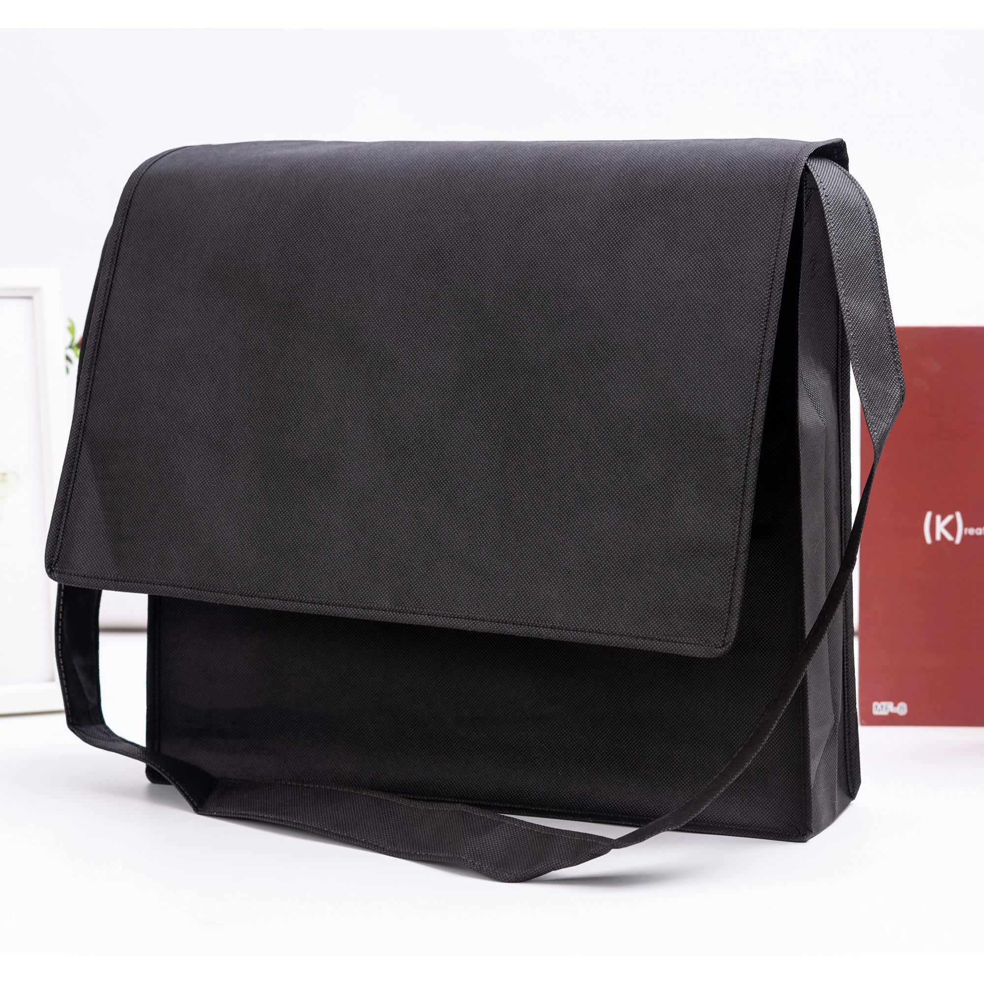 A black non-woven flap satchel with a shoulder strap and branding logo displayed. Non Woven Flap Satchel - Image 1