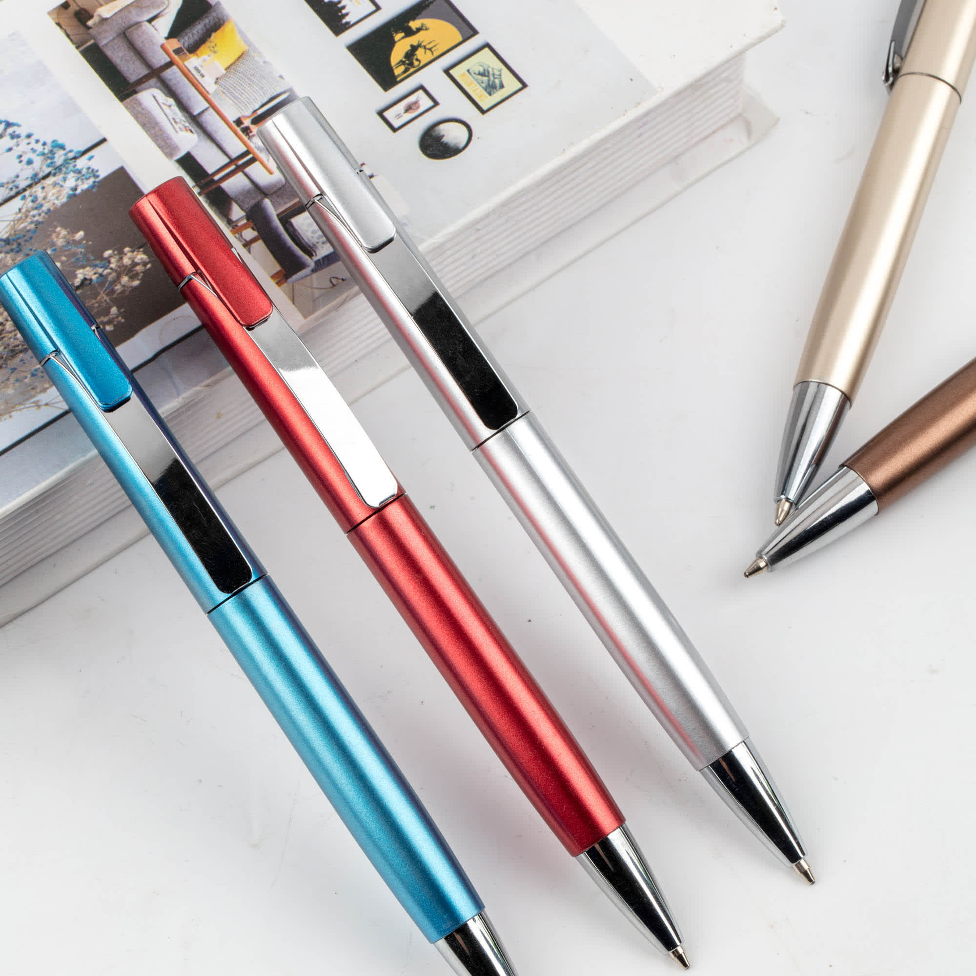 A set of metal pens in blue, red, silver, and bronze, resting beside a stack of books. Each pen has a branding logo. Shink - Image 1