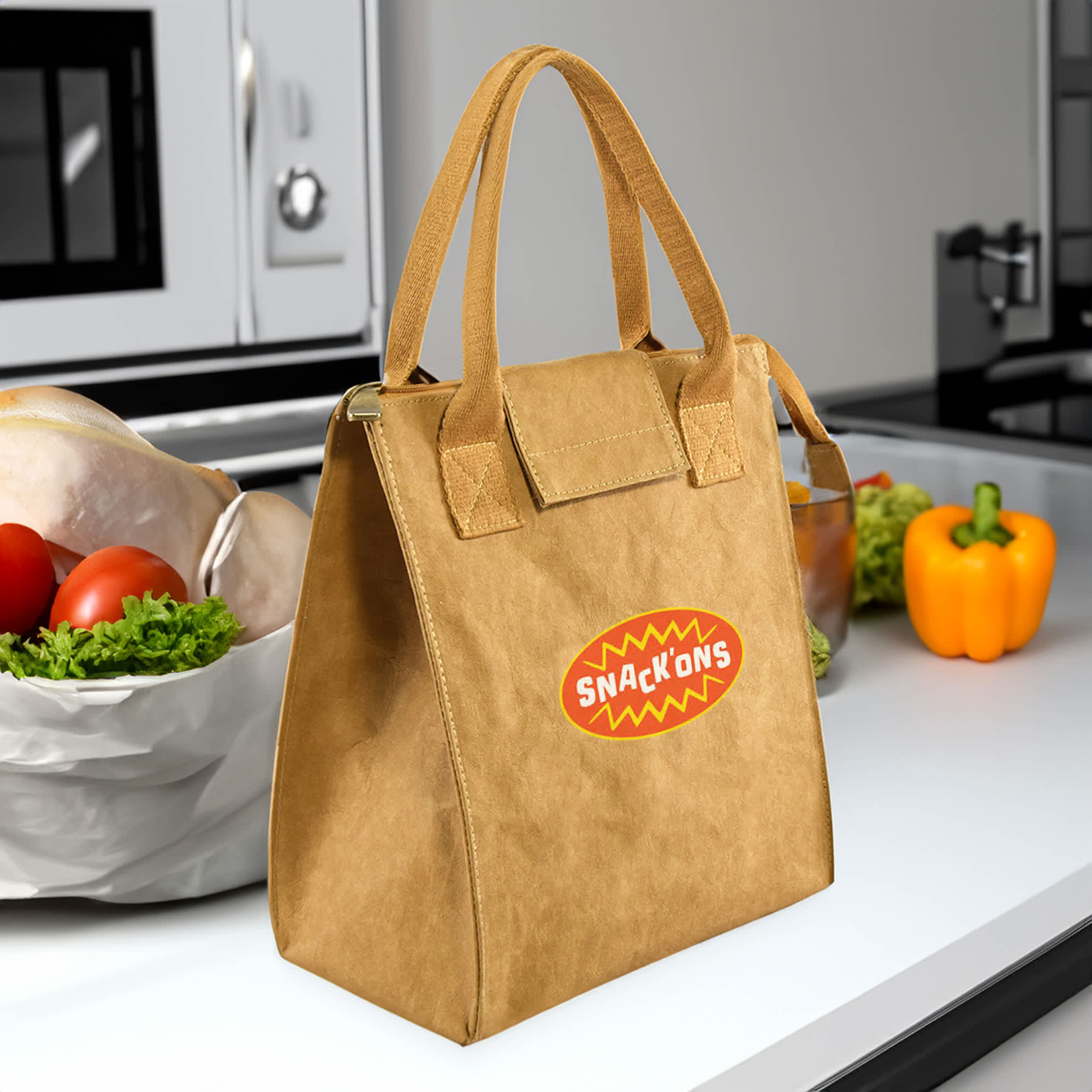 A brown lunch bag with handles, featuring a logo, placed on a countertop among fresh vegetables. Paper Cooler Lunch Bag - Image 1