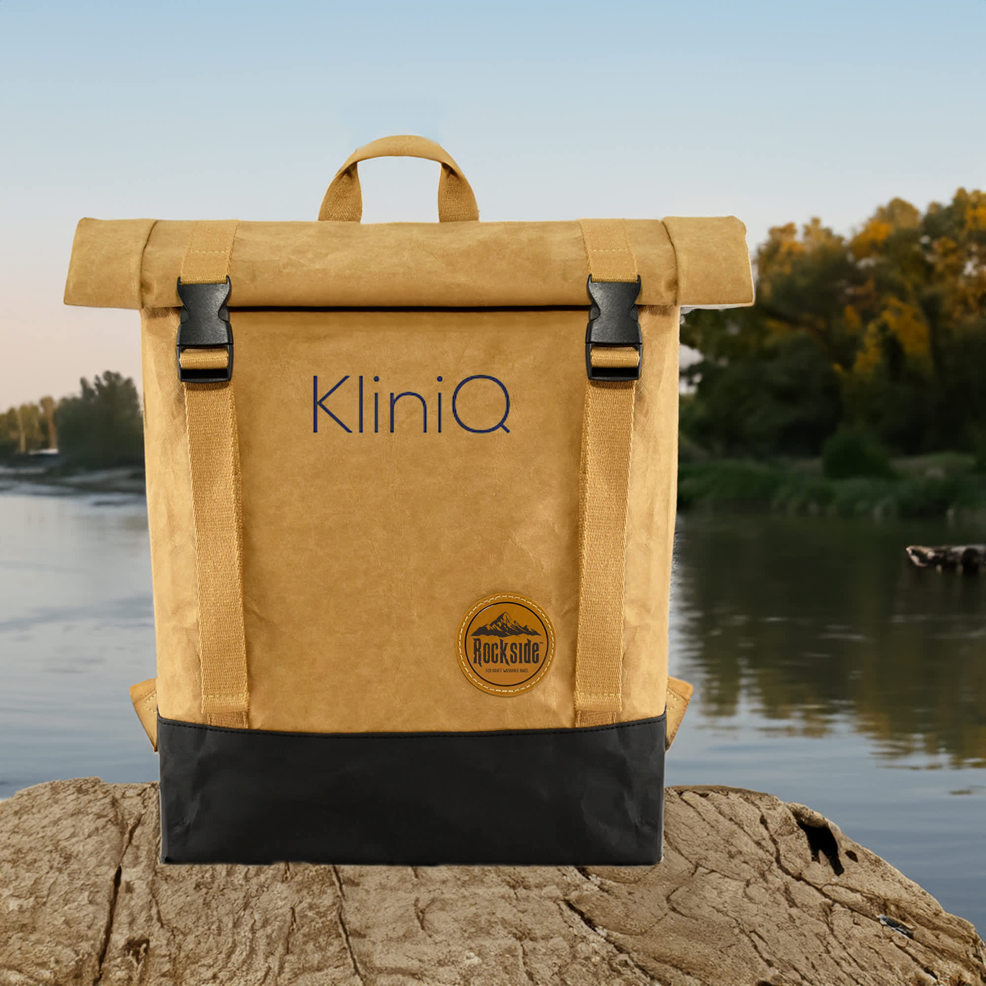 A kraft paper laptop backpack in tan and black, featuring straps and a logo, set against a scenic river backdrop. The Base Kraft Paper Laptop Backpack - Image 1