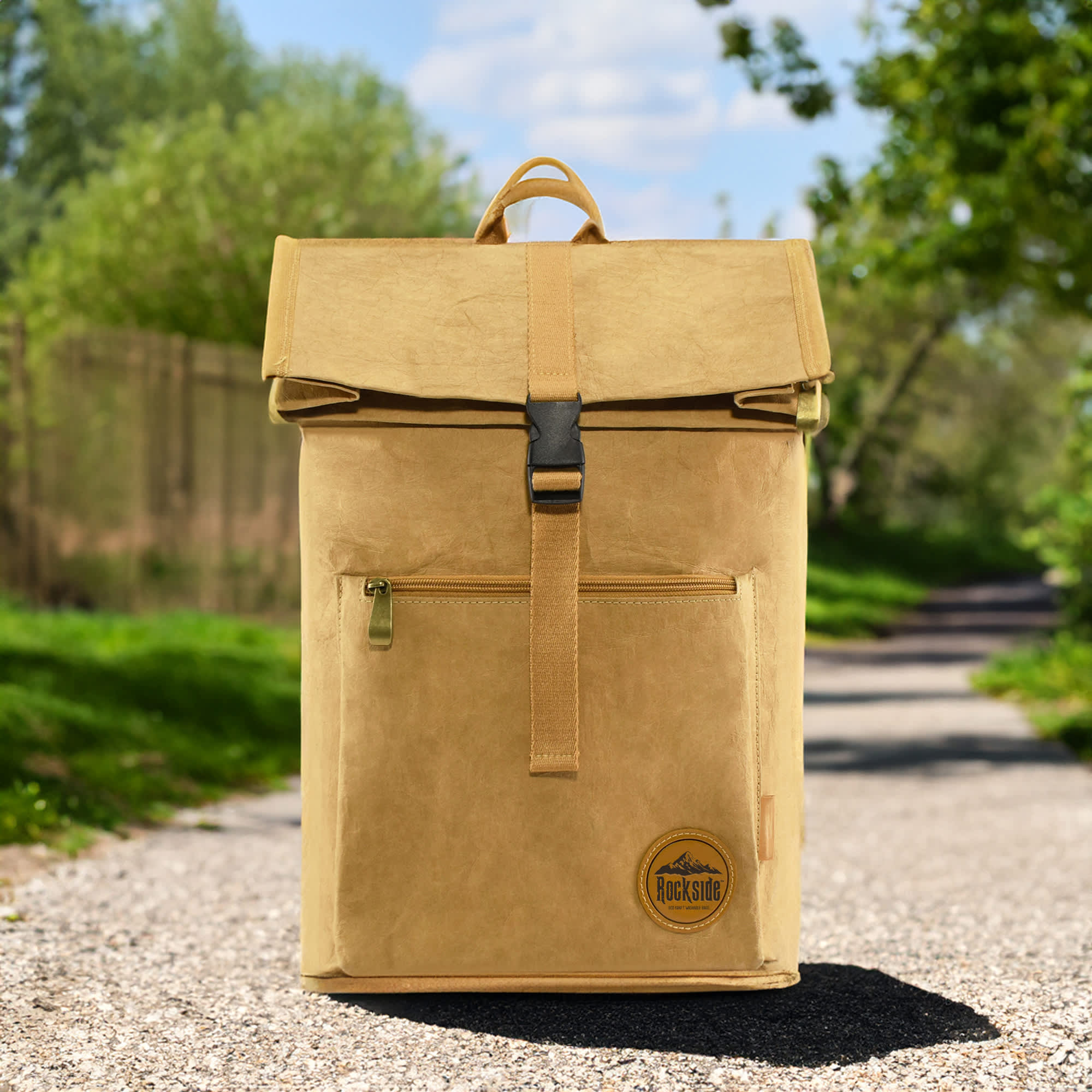 A yellow kraft paper laptop backpack with a fold-over top and front zip pocket, set against a scenic outdoor path. Somsonic Kraft Paper Laptop Backpack - Image 1