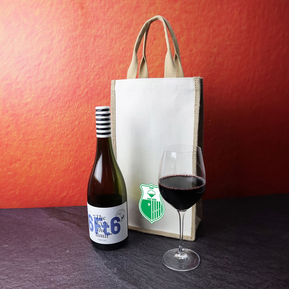 A jute double wine bag in natural colour alongside a bottle of wine and a glass of red wine. The bag has a logo. Jute Double Wine Bag - Image 1