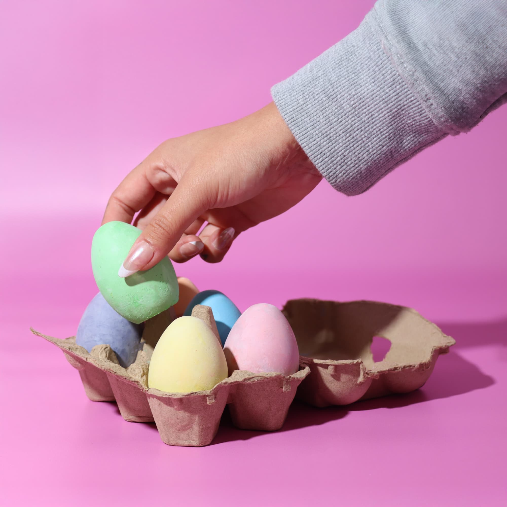 A hand reaching for a green pastel egg from a carton holding various pastel-coloured eggs on a pink background. Free Range Chalk - Image 1