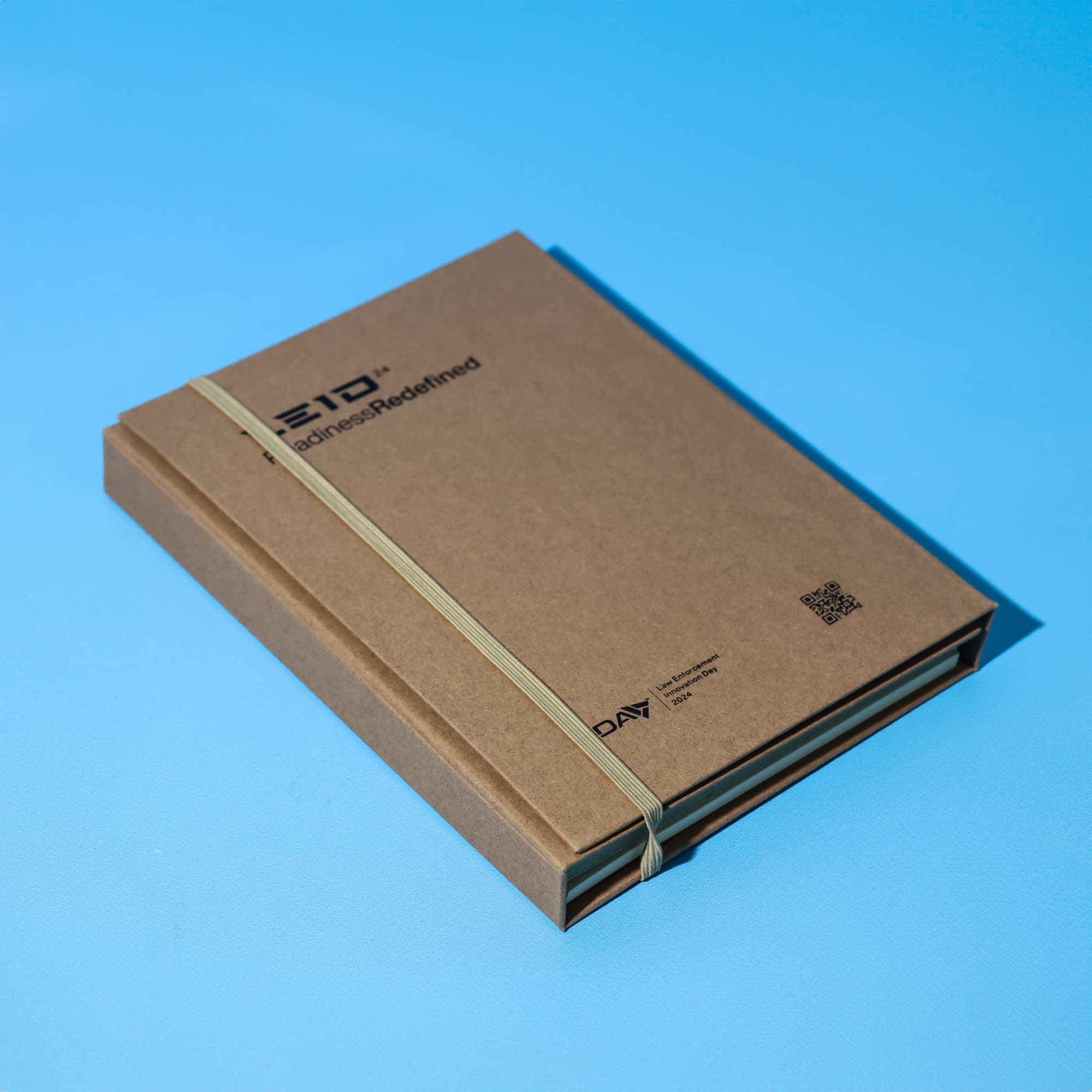 A brown A5 notepad secured with a rubber band, featuring a logo on the cover against a blue background. Essentials A5 Notepad - Image 1