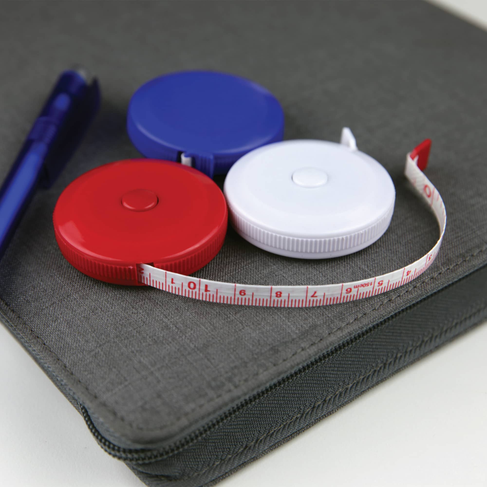 A compact tape measure in red, blue, and white, lying next to a pen on a grey surface. It has a logo. Compact Tape Measure - Image 1