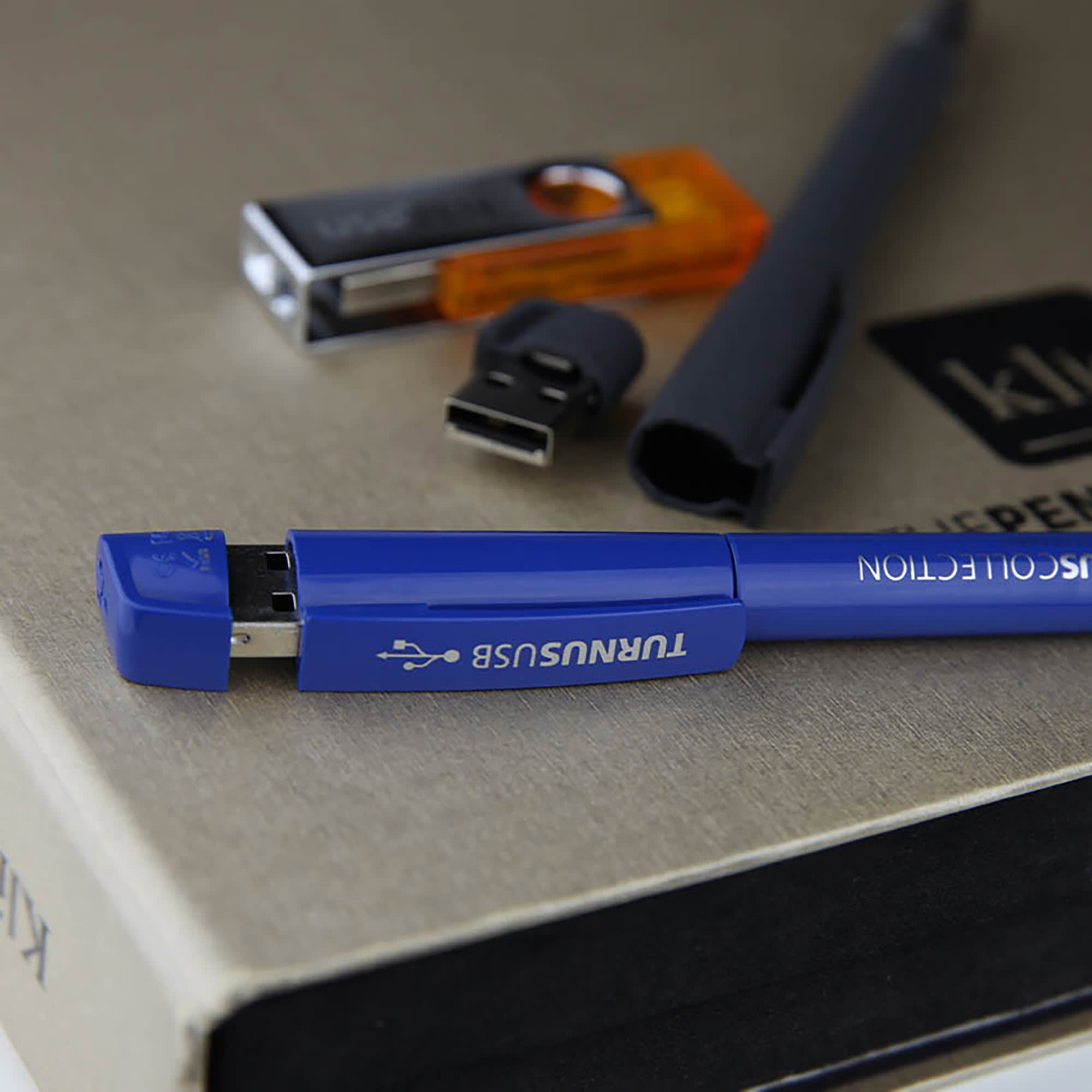 A blue USB pen with a metal tip beside other flash drives on a textured surface. USB Pen 16GB Metal Tip - Image 1
