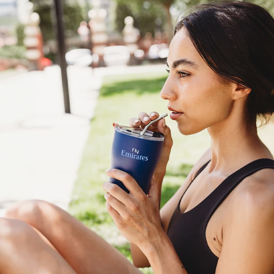 A drink bottle in navy blue with a stainless steel straw and a logo, held by a woman outdoors. Wanderer 354ml Tumbler - Image 1
