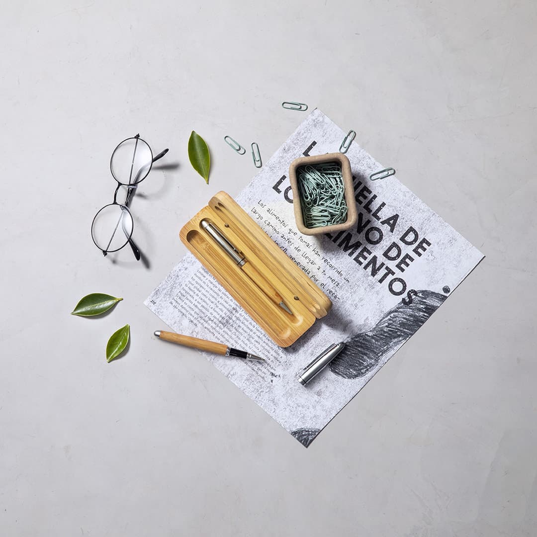 A wooden pen case with a pen, paperclips, and glasses, surrounded by leaves and a piece of paper. Notium - Image 1