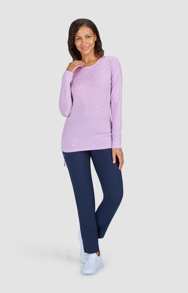 A woman in a lilac long-sleeve top and navy trousers stands with one hand on her hip, smiling. The outfit features a logo. Tessa Tee (Print) - Image 1