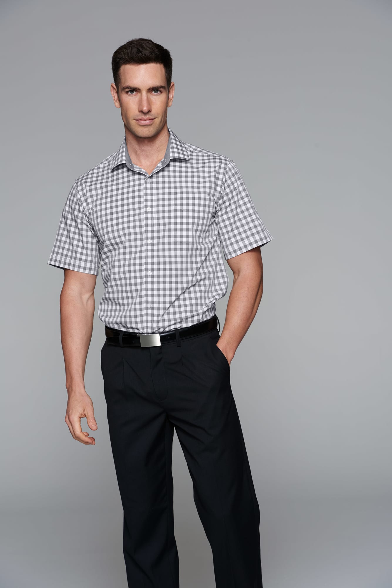 A man models a short sleeve, button-up shirt in a grey check pattern, paired with dark trousers. The shirt has a logo. DEVONPORT MENS SHIRT SHORT SLEEVE RUNOUT - Image 1