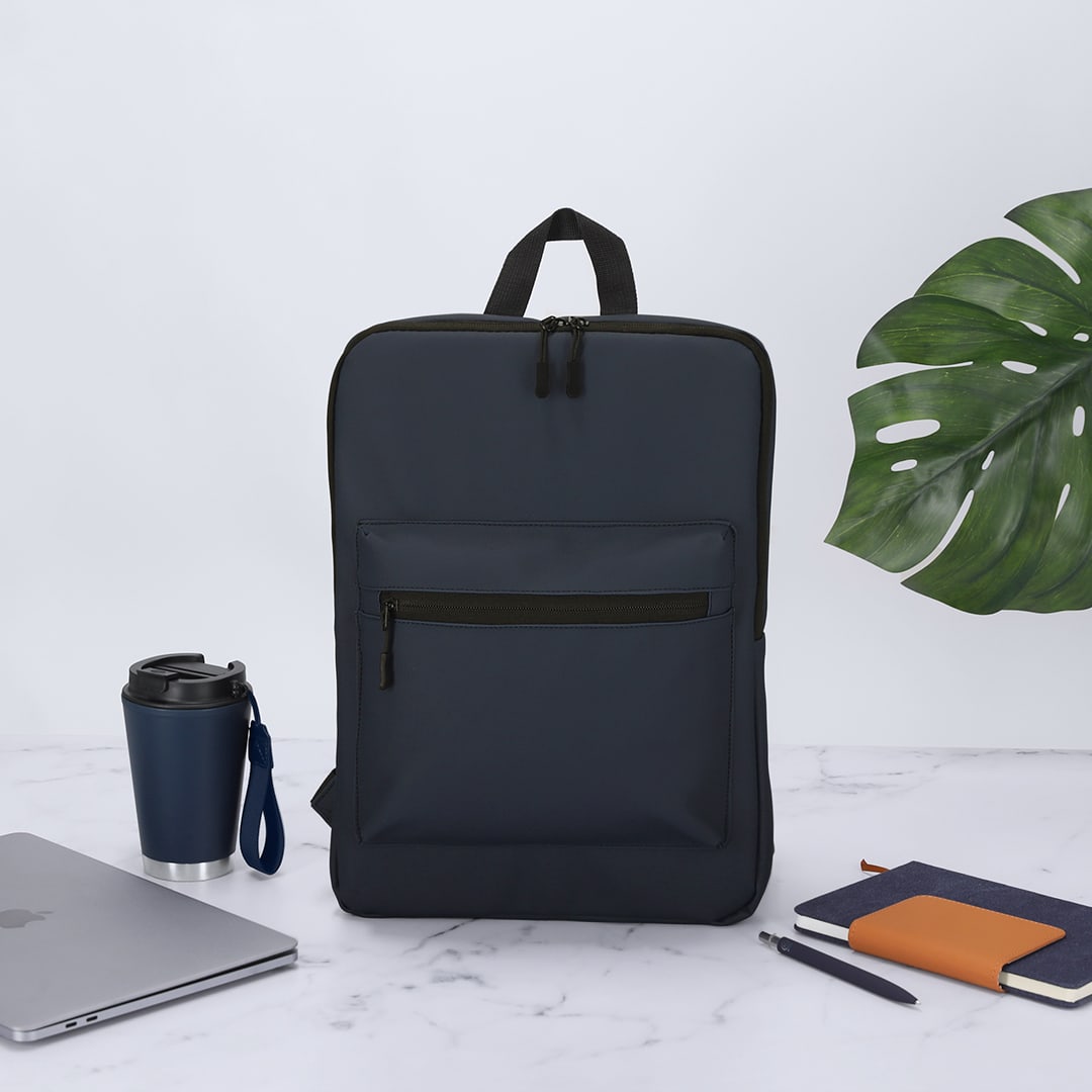 A navy backpack with a front pocket, placed beside a drink bottle, laptop, notebook, and a plant leaf. Yilmir Backpack - Image 1