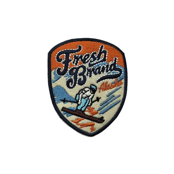 A custom embroidery patch featuring a snowy mountain scene with a skier, predominantly in blue, orange, and beige. Custom Embroidery Patch - Image 1