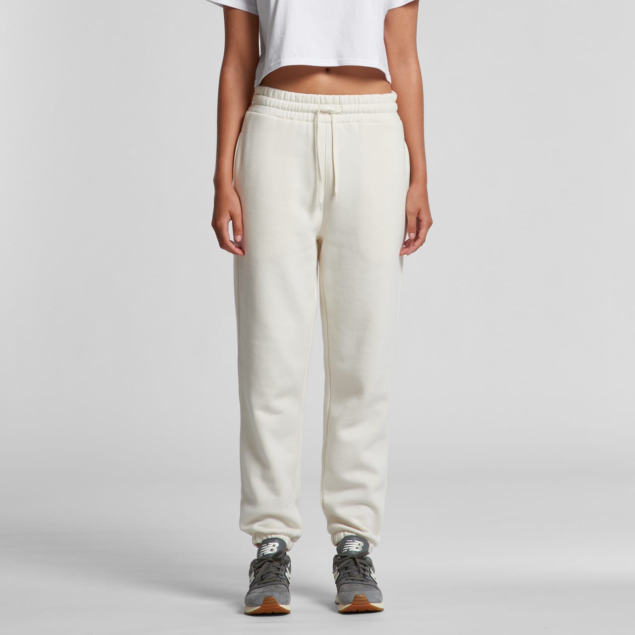 A pair of cream track pants with a drawstring waist and a logo, styled with a cropped top and grey sneakers. Wo's Stencil Track Pants - Image 1