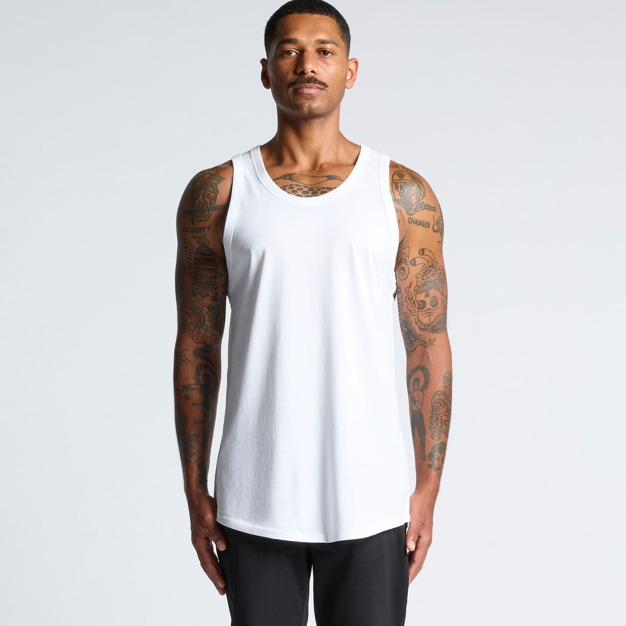 A man wearing a white sleeveless singlet, featuring tattoos on his arms, standing against a light background. Authentic Singlet - Image 1