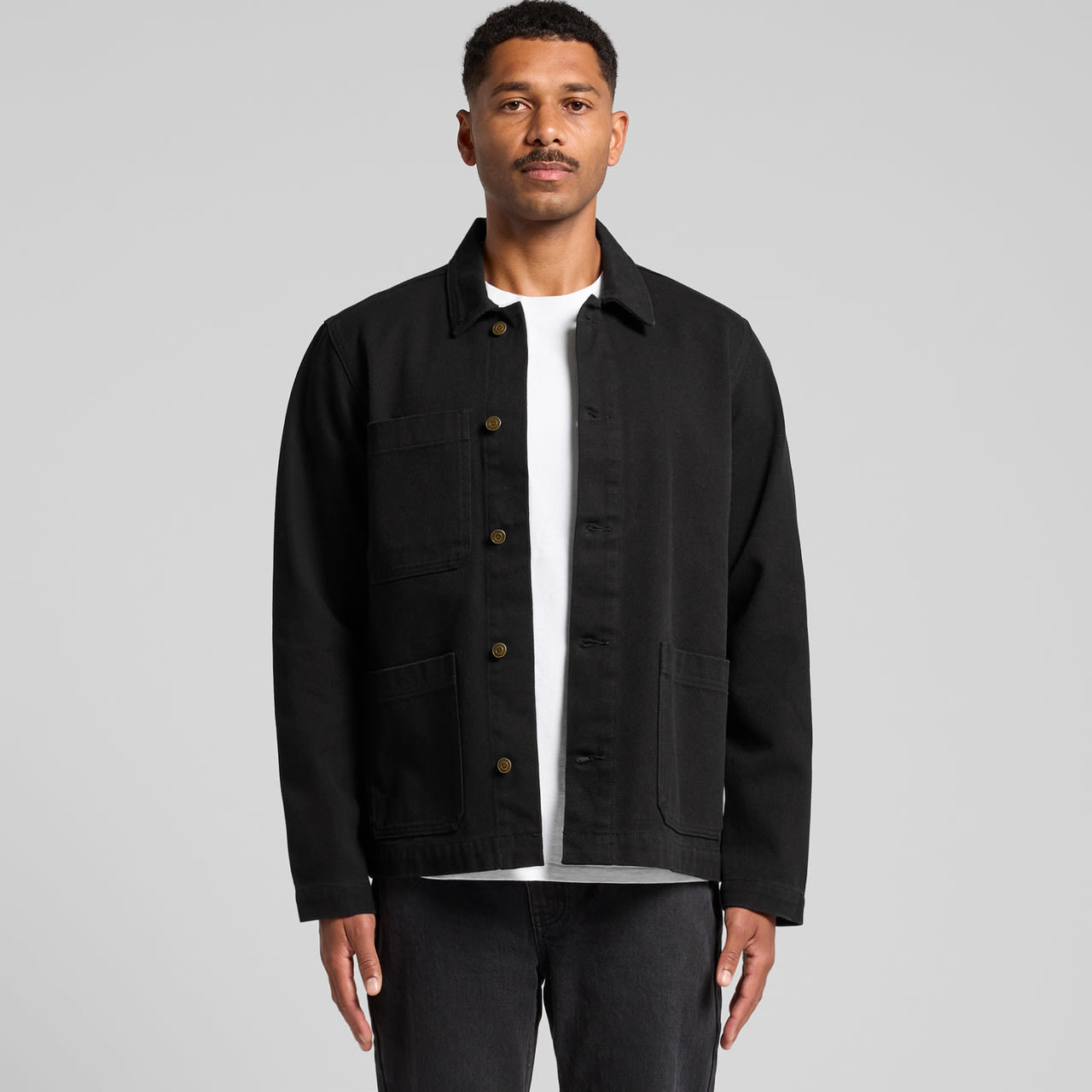 A man wearing a black canvas chore jacket with button closure and multiple pockets. The background is plain grey. Canvas Chore Jacket - Image 1